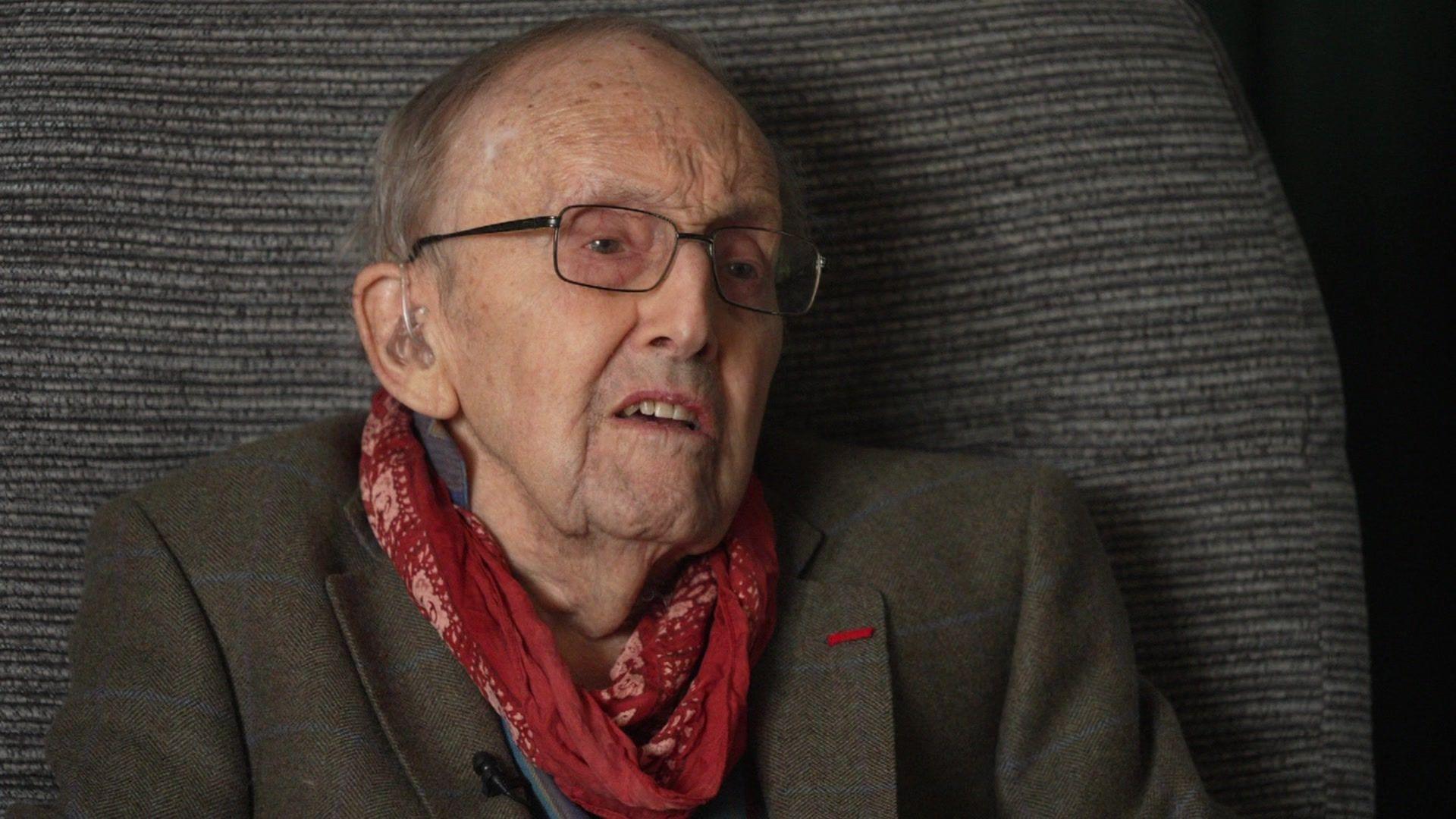 Cecil Newton, an elderly white male wearing thing black framed glasses and a hearing aid, with a red cravat and green tweed blazer sitting in his armchair