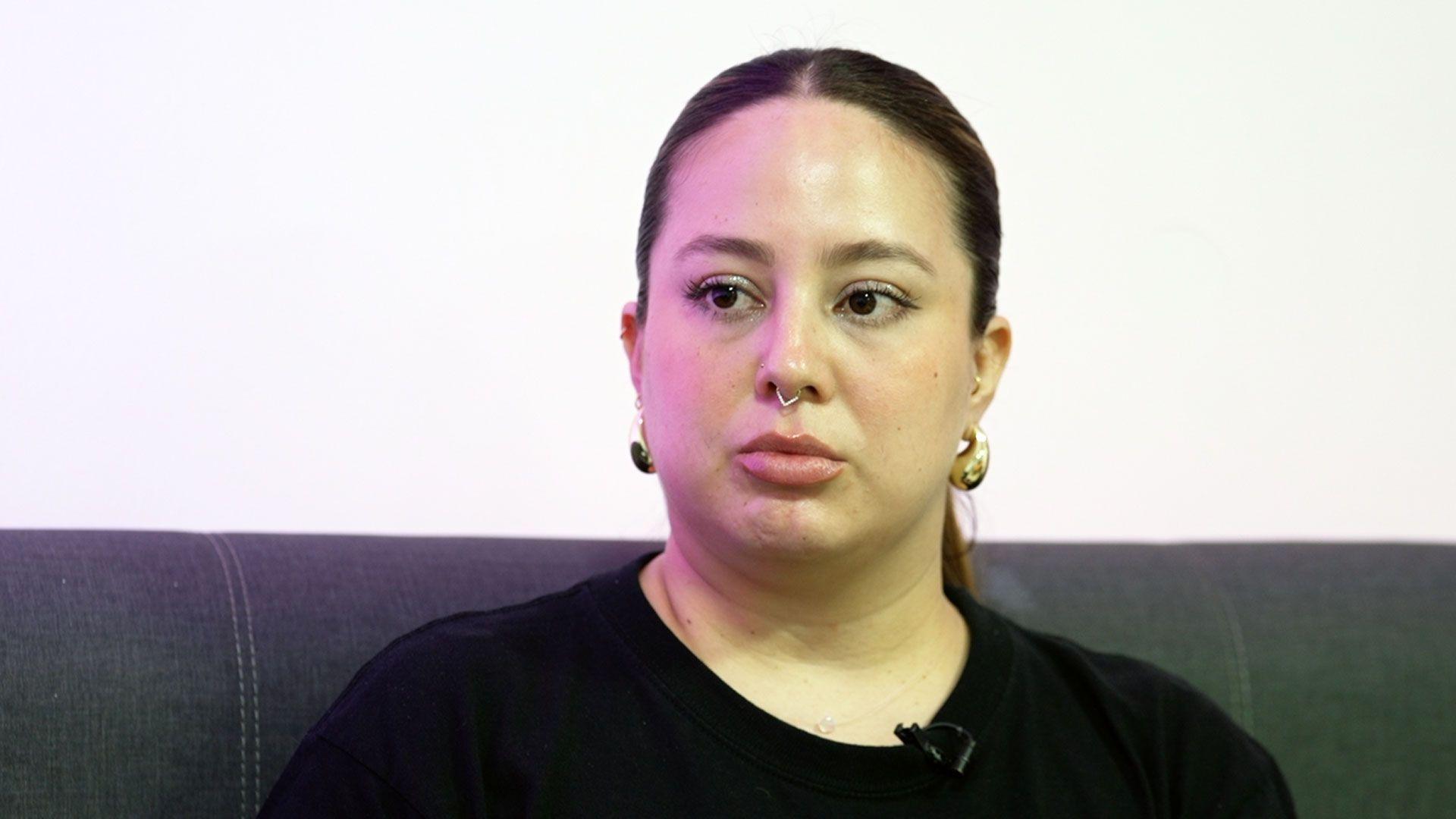 A young woman with her dark hair tied back, wearing gold earrings sits on a grey sofa.