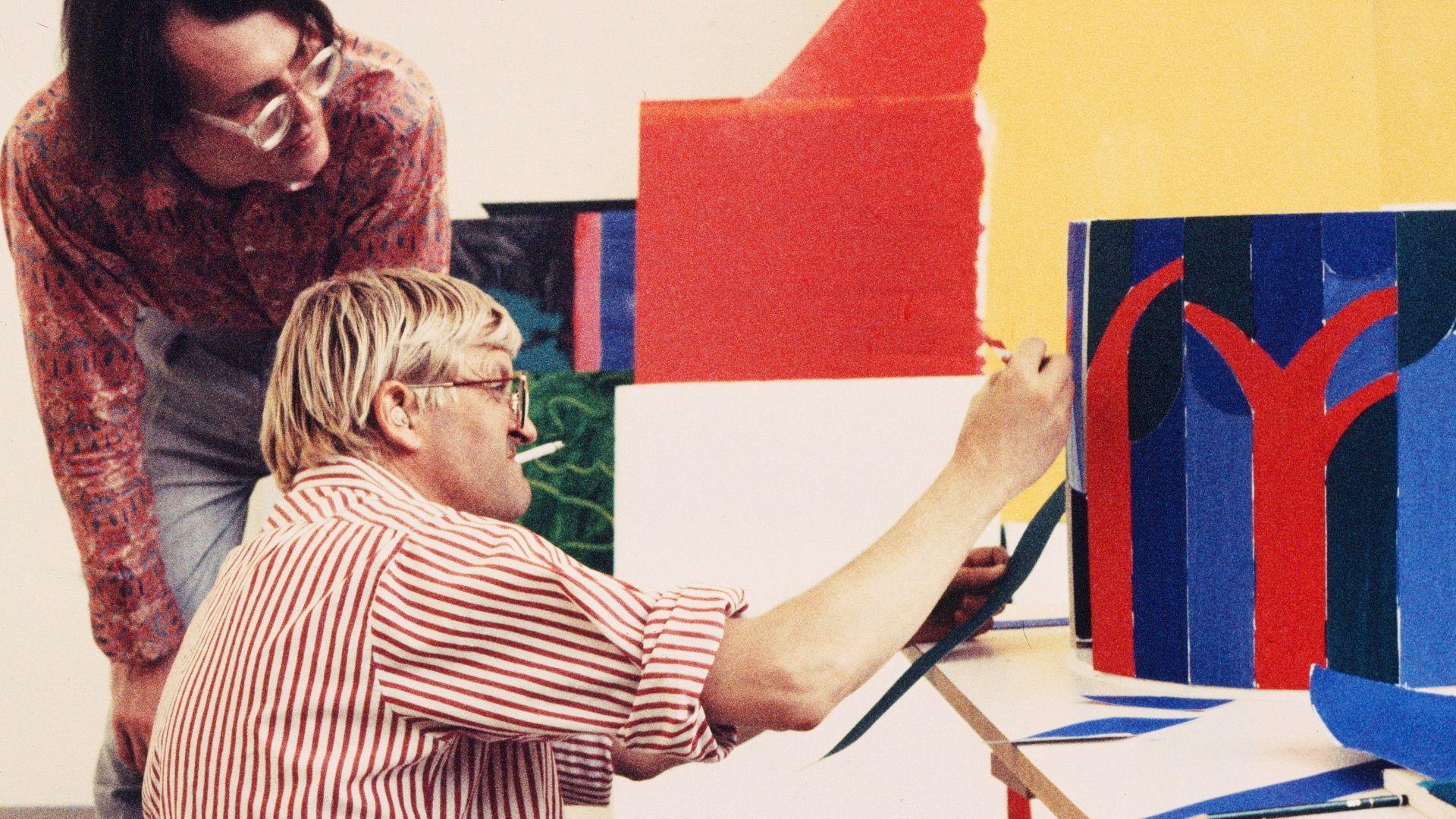 A young David Hockney smokes a cigarette as he paints a blue and red structure with someone peering over his shoulder. 