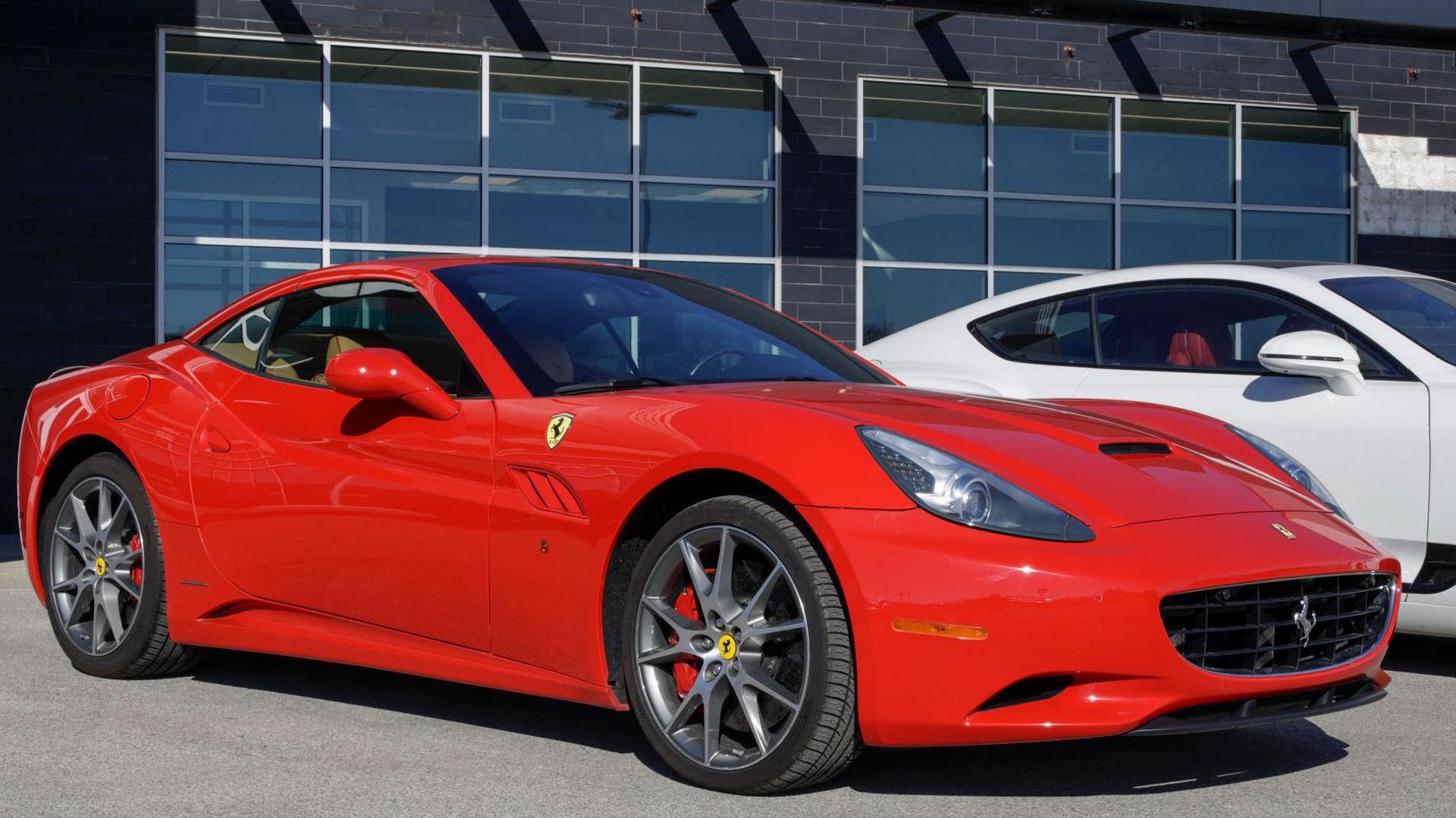 A red Ferrari sports car is seen on a forecourt outside a dealership. There is a white model partially visible behind it