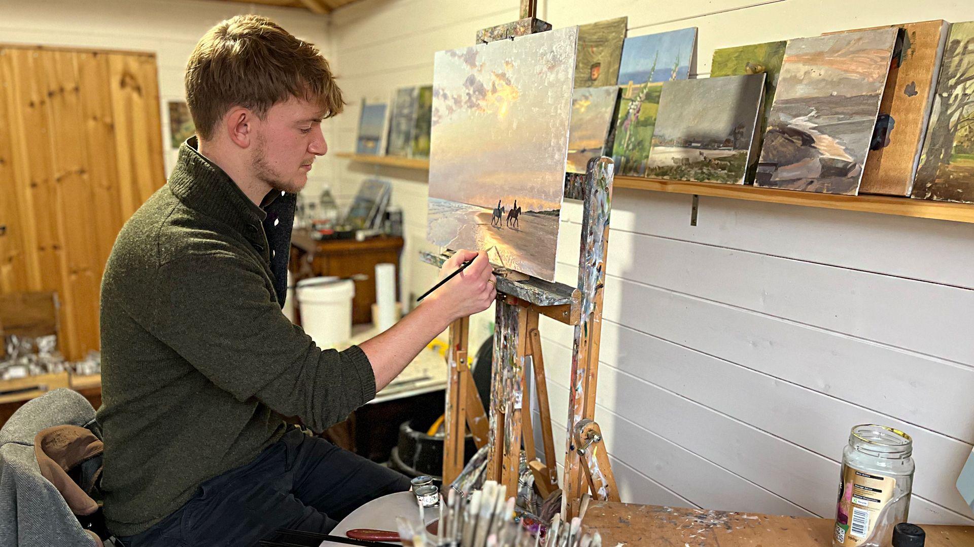 Kieron Williamson in his art studio, adding touches to a painting that shows two horses being ridden on a beach. He is wearing a green jumper and black trousers. On a shelf are various paintings he has produced. 