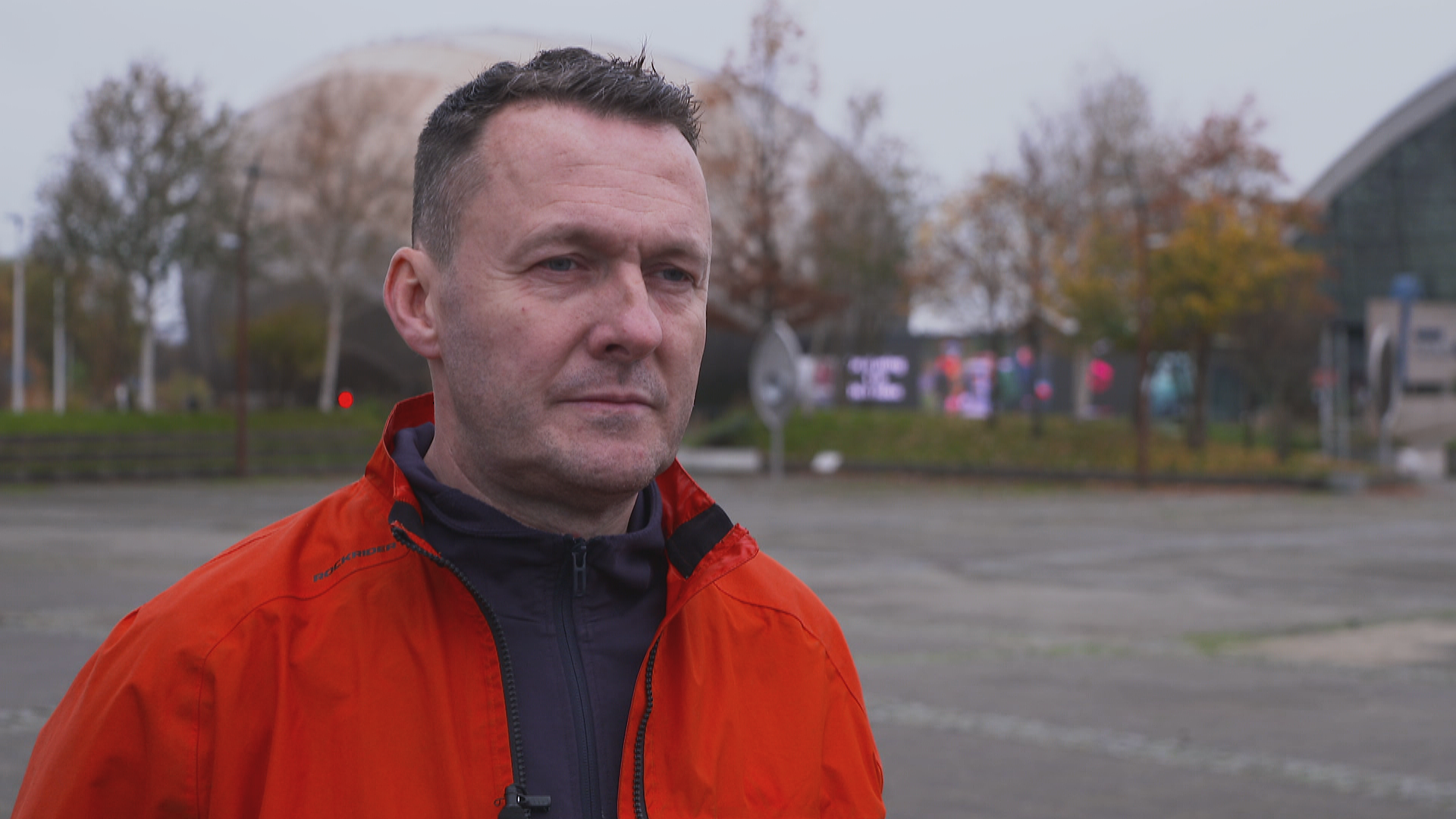 Russell Findlay wearing a red waterproof jacket with a dark coloured top underneath, standing in front of the SEC in Glasgow