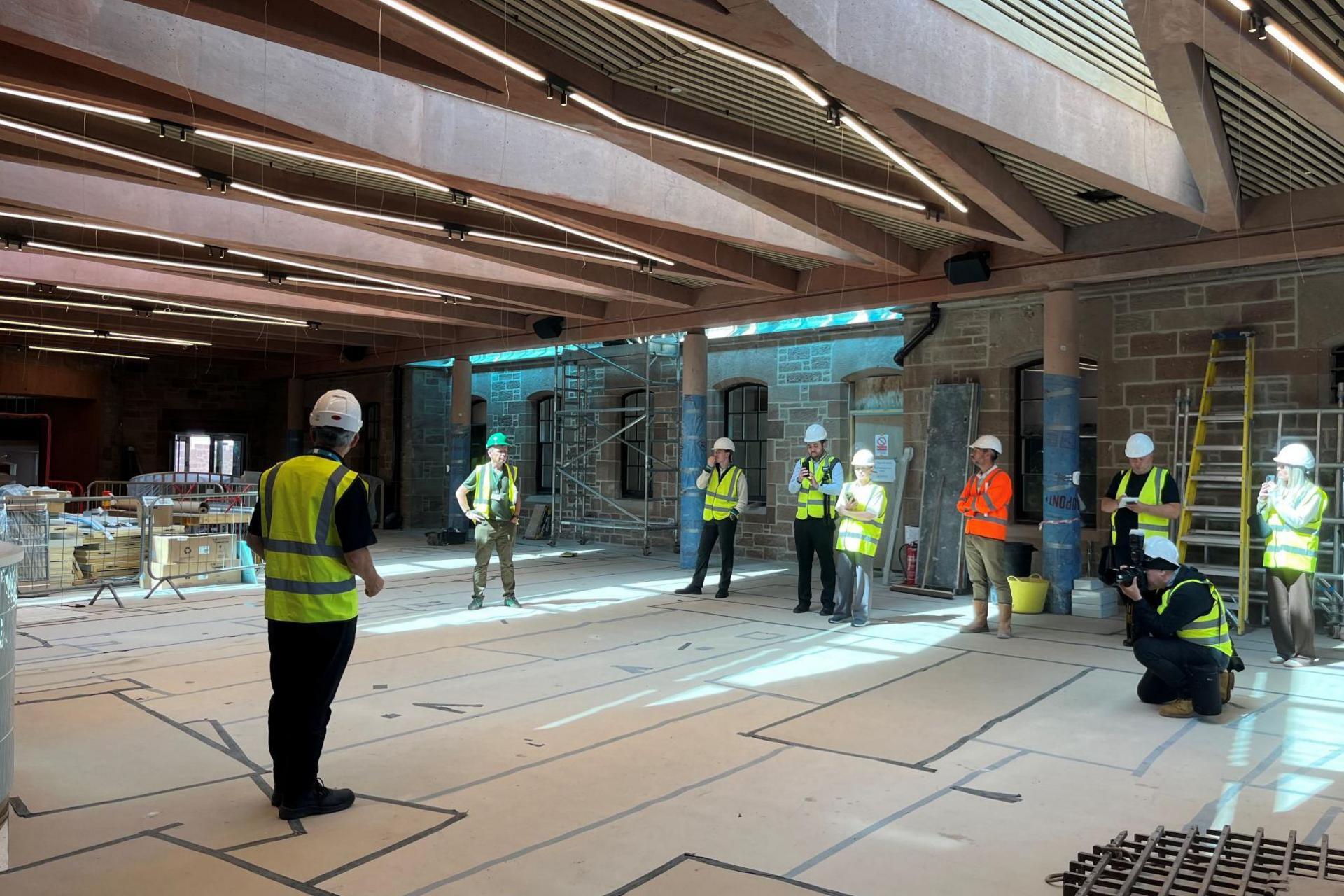 Nine people wearing high visibility waistcoats and white hard hats are inside a large room. One of the people is crouched down and taking a photograph.