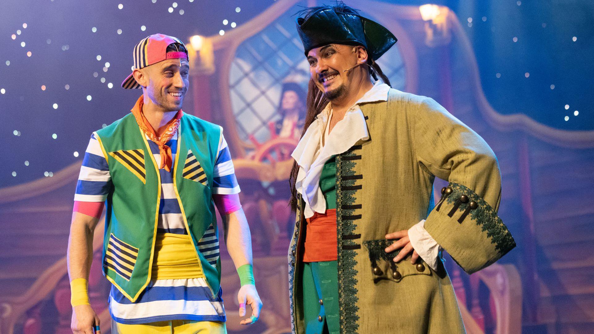 Daniel Bell on stage with another performer in the panto Treasure Island. He is on the left and is wearing a cap, which he has on backwards, a waistcoat, striped top and trousers. His fellow performer, on the right, is dressed as the pirate Captain Hook. He has a black hat, long hair, a beard, a jacket, white shirt, red cummerbund and a green jacket. The set behind them is out of focus, but it appears to be a pirate ship.