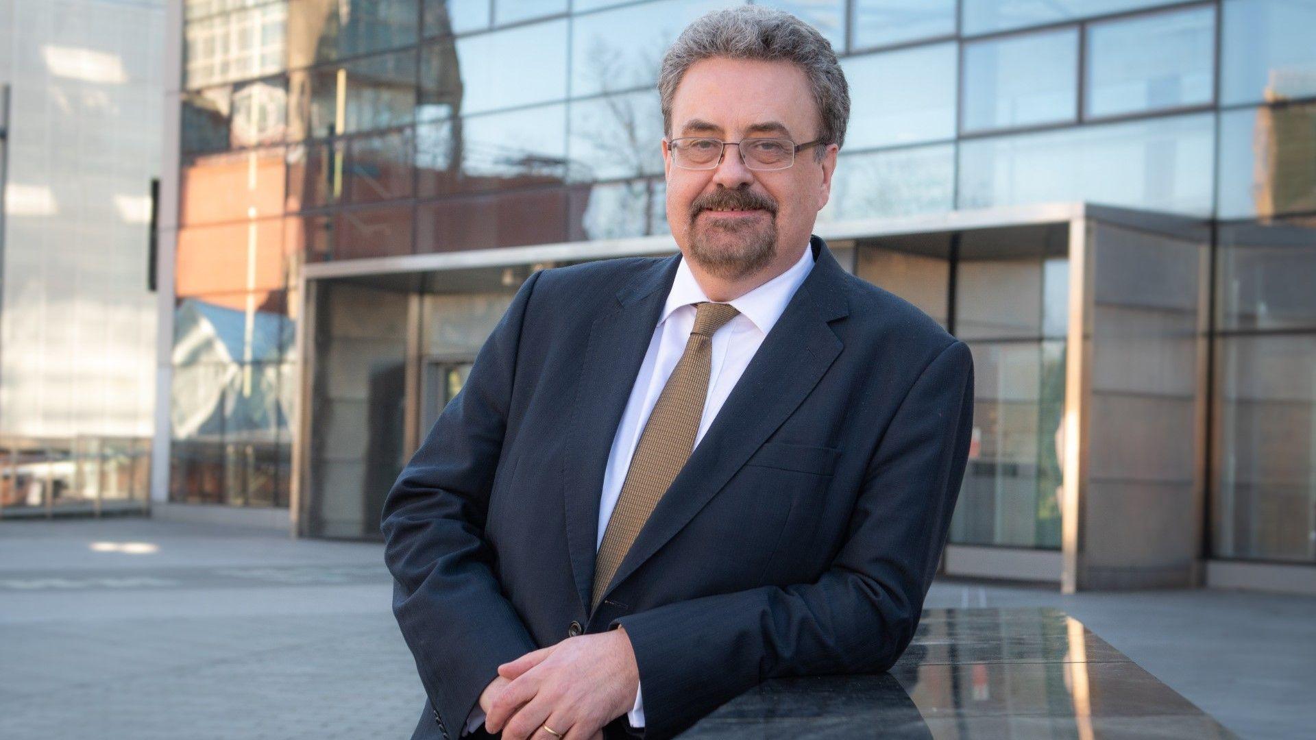 A man wearing a blue suit and brown tie, with glasses, leans against a wall outside a university building