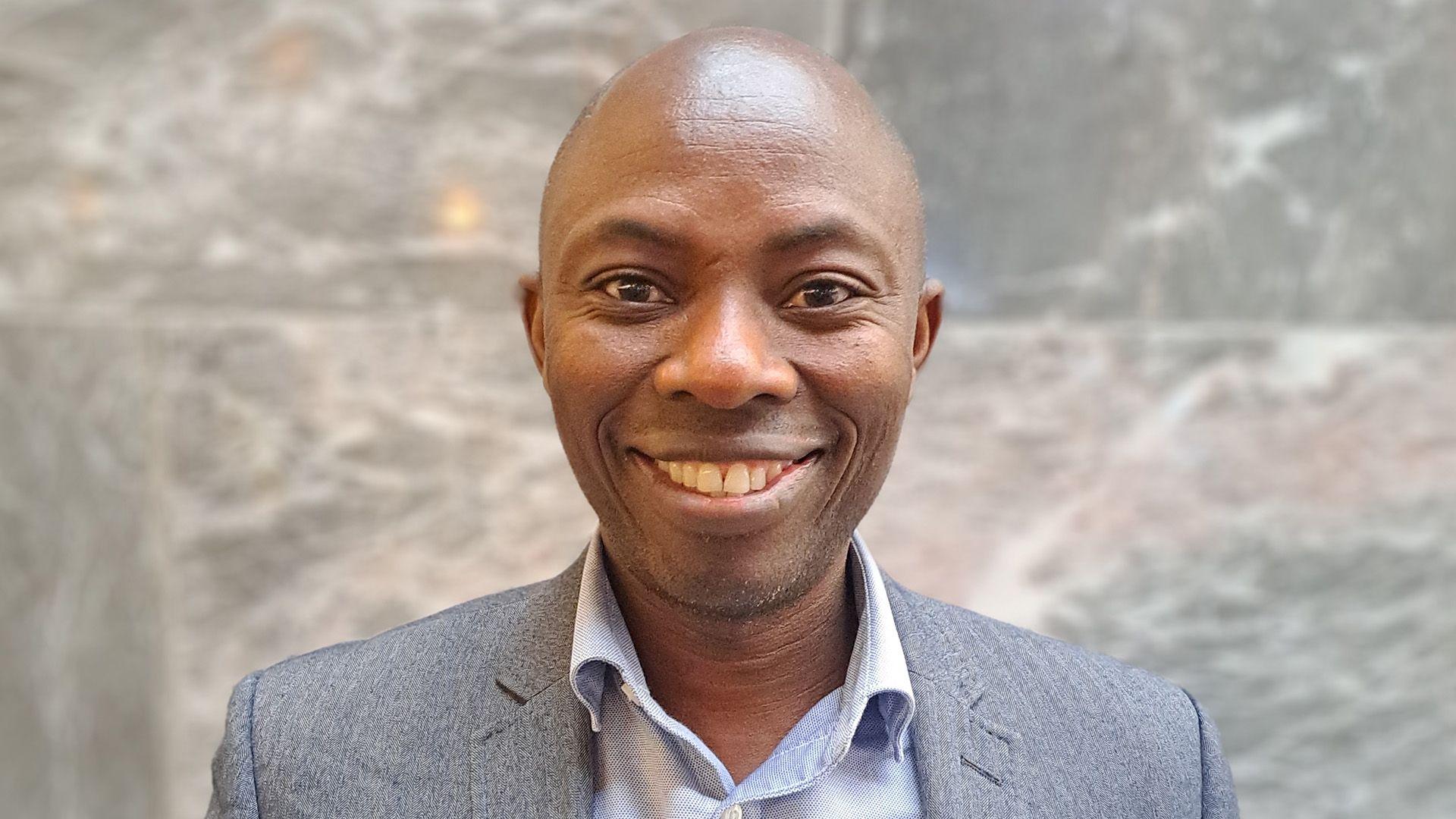 head and shoulders of man, looking at camera, bald and clean shaven, smiling, wearing grey suit