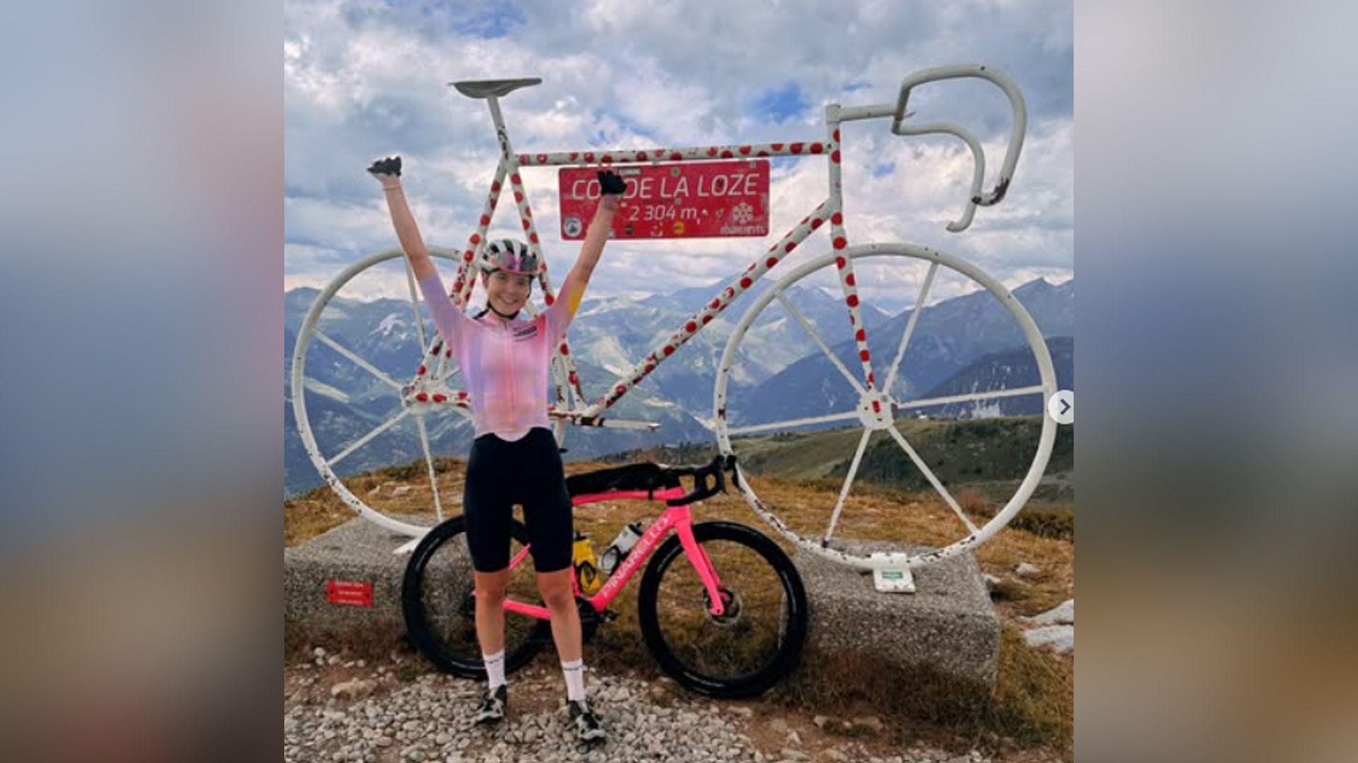 A woman stands with arms aloft by a parked pink mountain bike, next to a large sculpture of a bike in white metal tubing, decorated with red spots and with a sign reading "Col de la Loz - 2,304m" 
