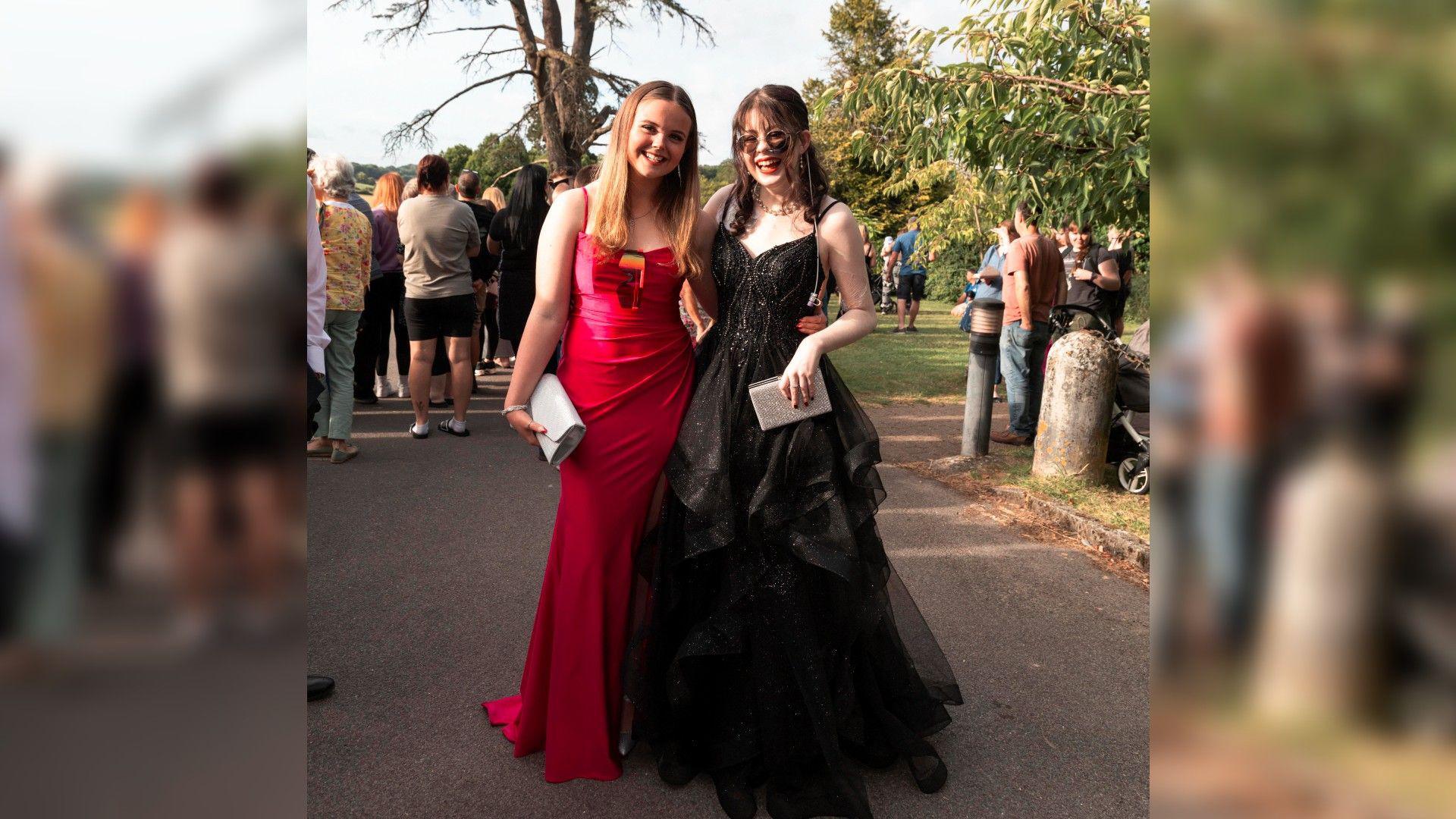 Ellie in a black dress smiling and looking at the camera stood next to her friend in a red dress. Both girls look very happy and are stood outside on their prom day.