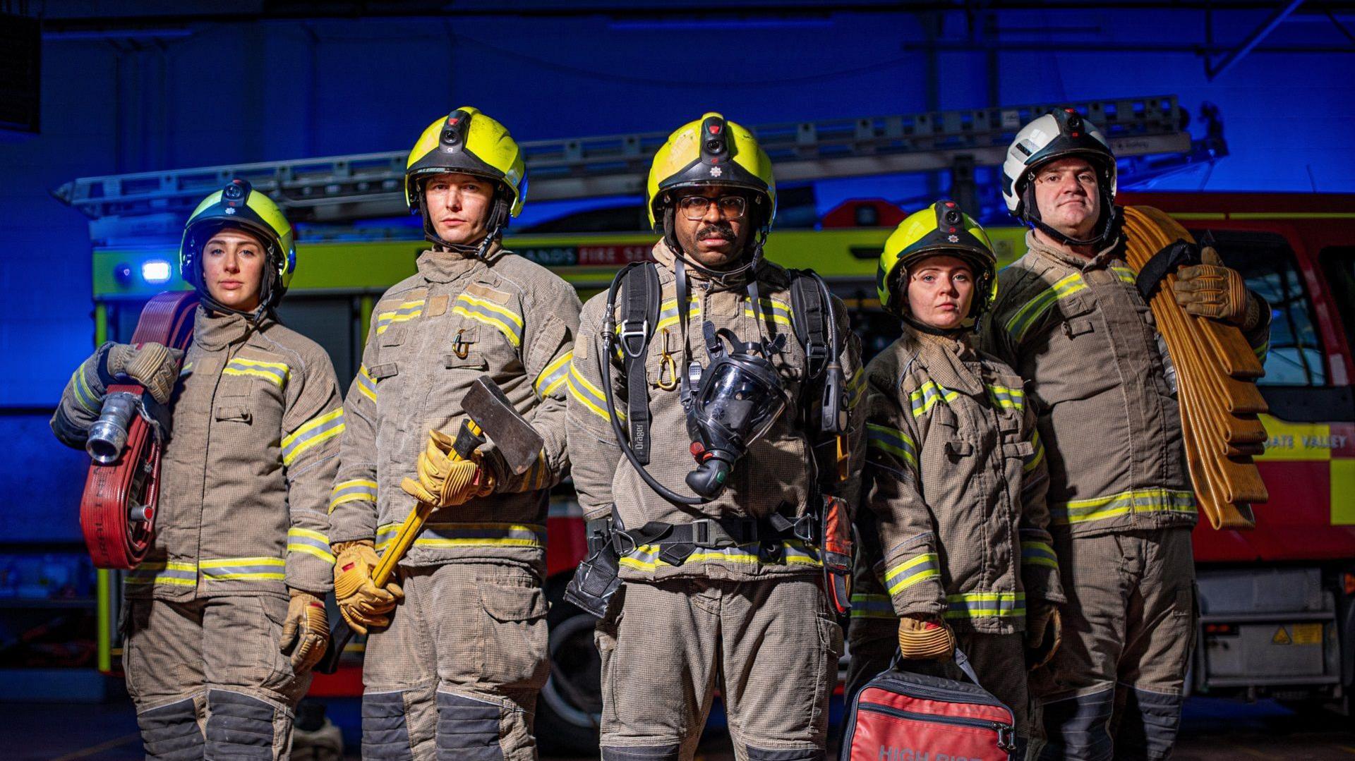 Five firefighters in brown fire suits with green reflective strips are standing in a line, in front of a fire engine. They are all wearing helmets. Each are holding a piece of kit, including hose reels and an axe.