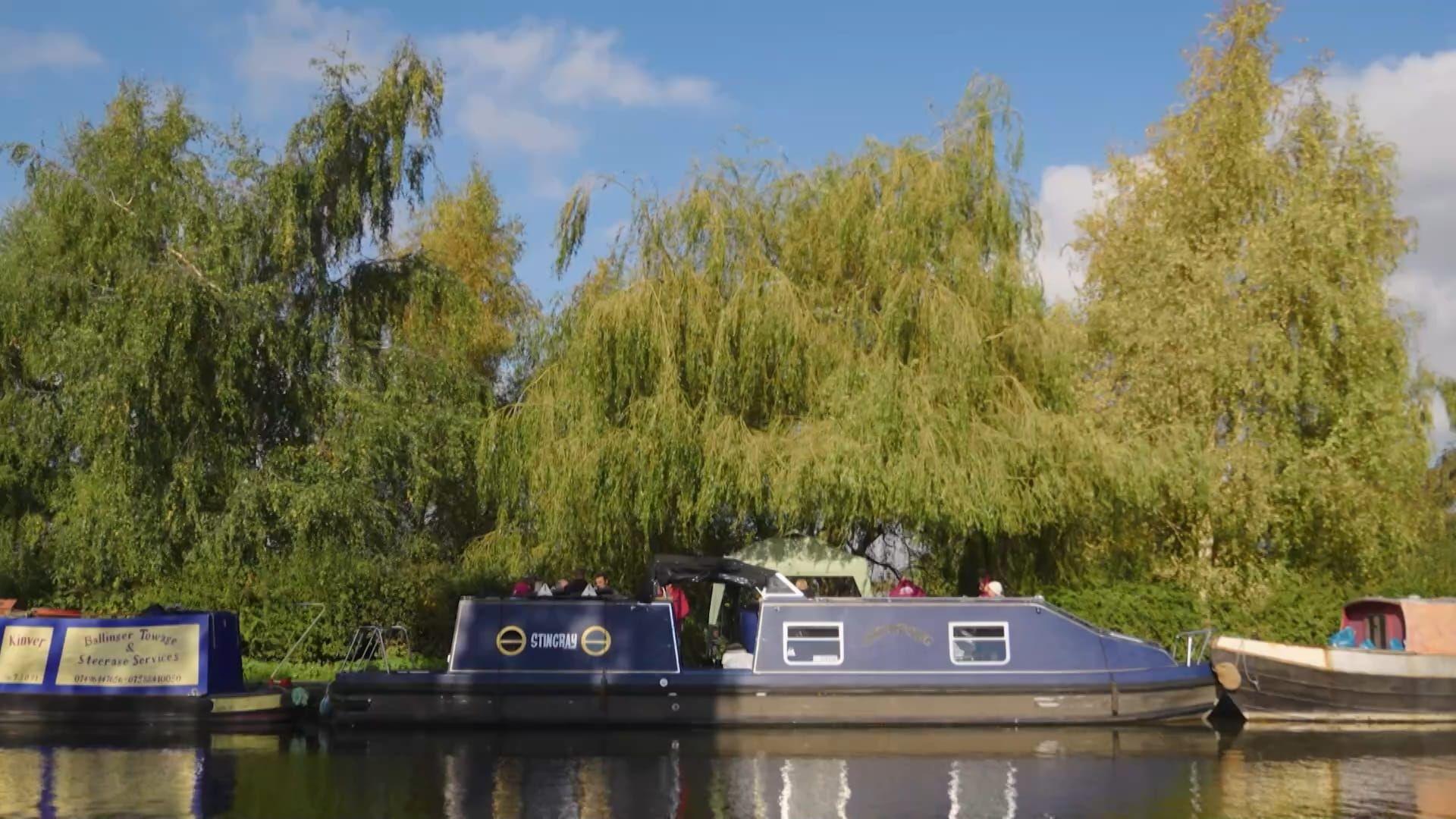 A side show of the vessel Stingray as it sits, moored to a canal bank beside some trees