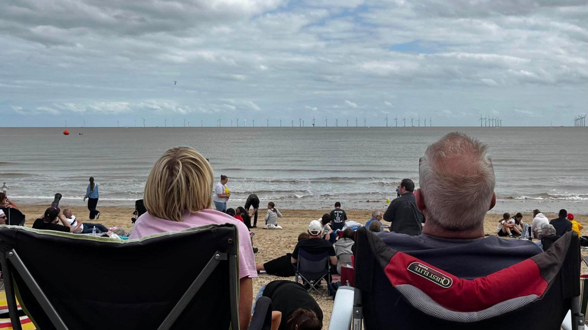 A picture taken from behind a woman and a man who are sitting on camping chairs on the beach. They are looking above the sea at the sky. A plane, which cannot be seen in the picture, is in flight.
