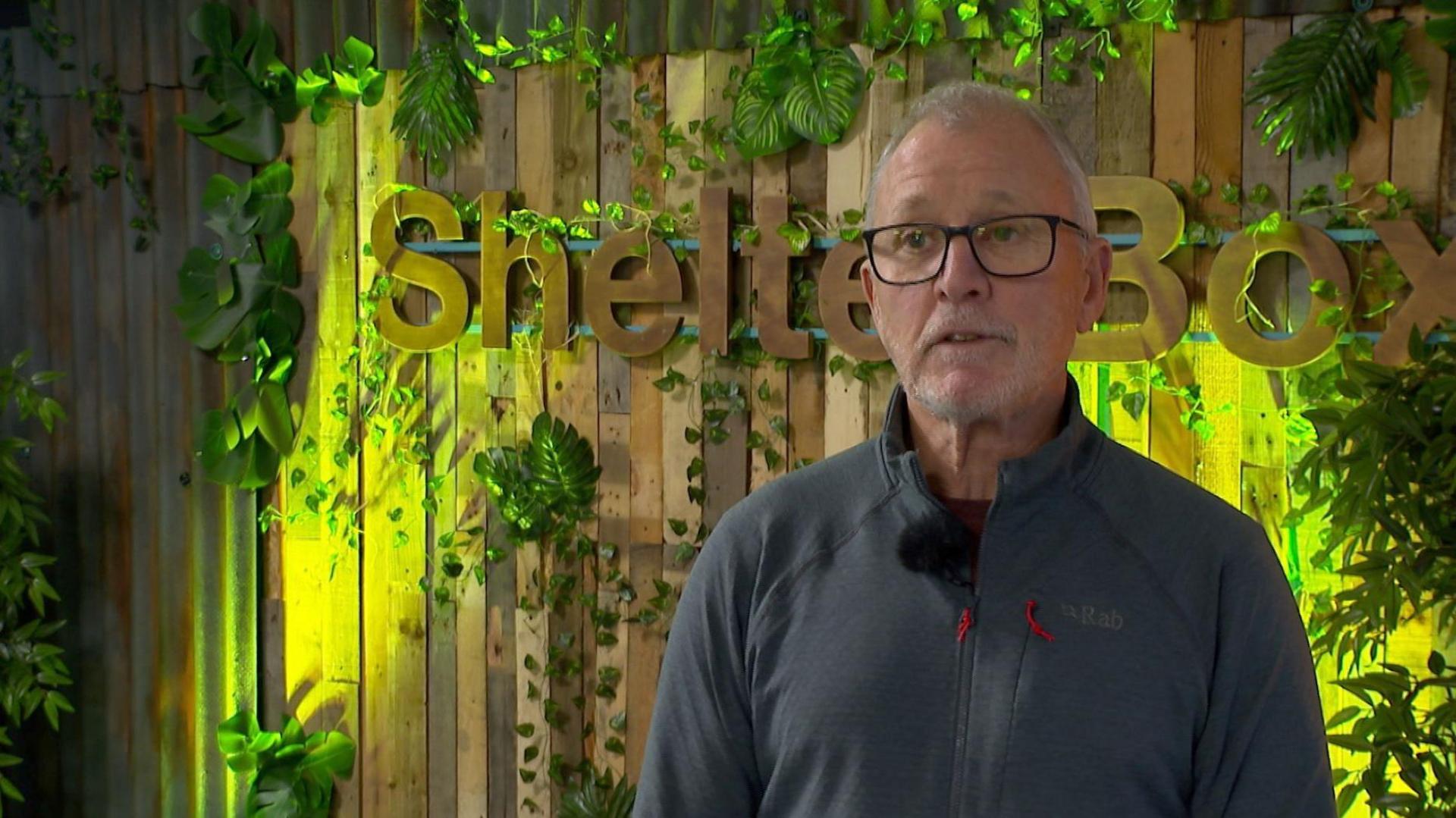 A man with short grey hair, glasses and a blue zipped-up top stands in front of a wooden, illuminated Shelterbox logo surrounded by plants.