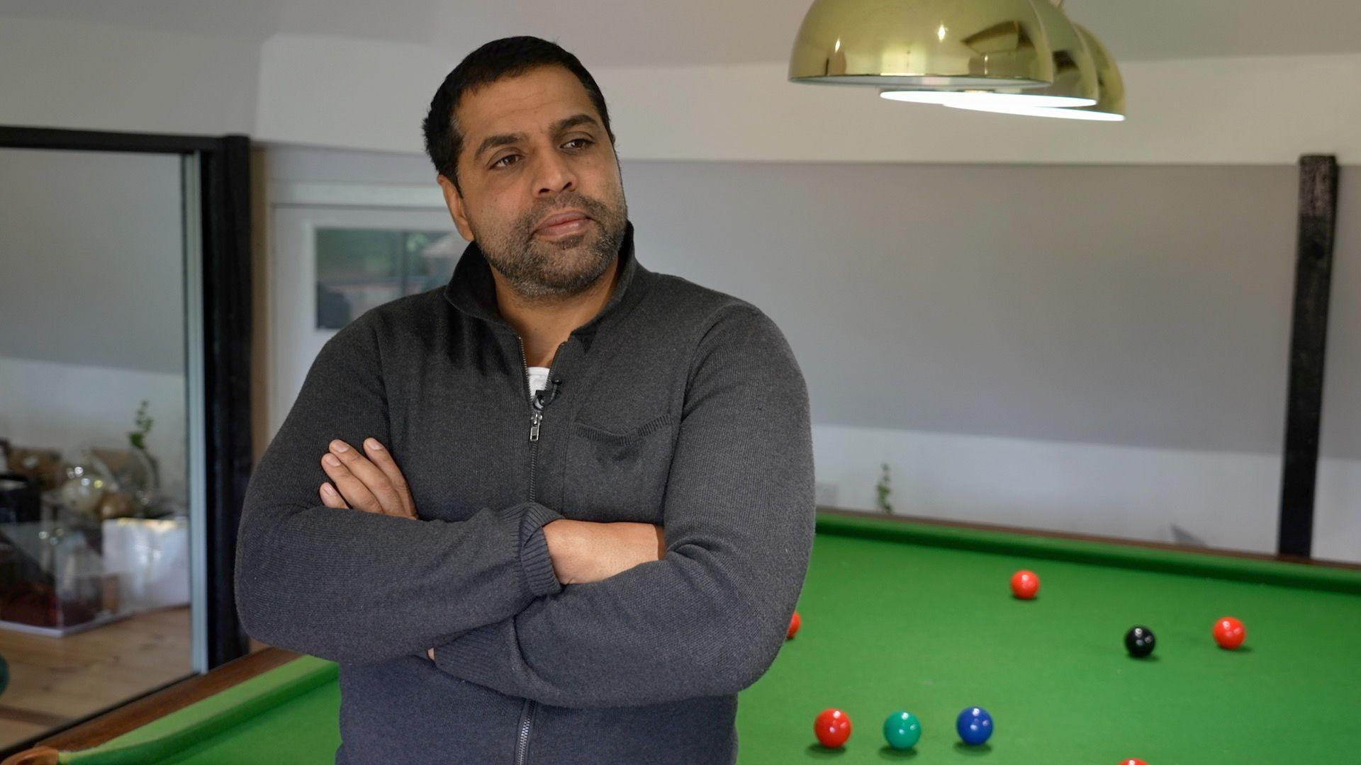 A man in a dark grey zip up jumper is leaning against a pool table with his arms crossed across his chest and is looking towards the right of the camera.