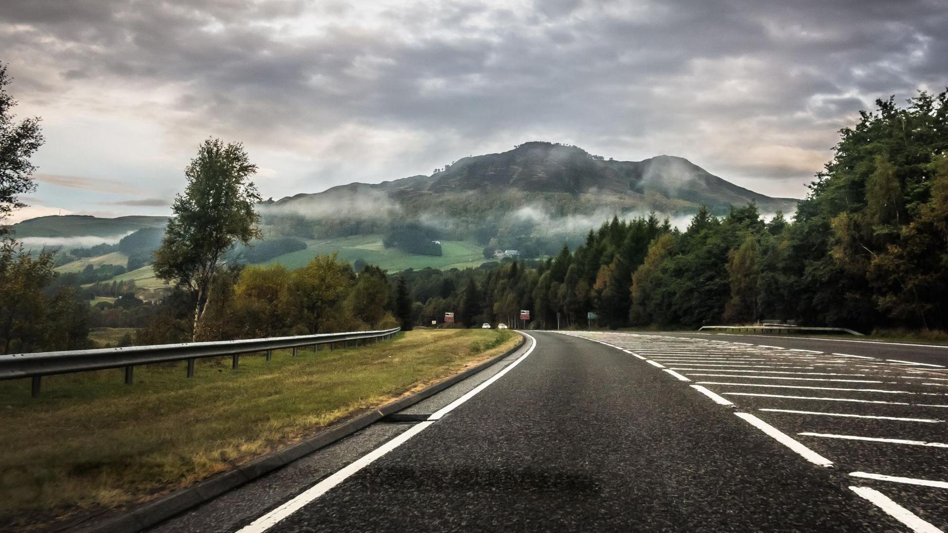 A crash barrier runs along the side of the road, which has a thick white lines on both sides and chevrons in the middle. Trees also line the side of the road and there is a hill in the distance.