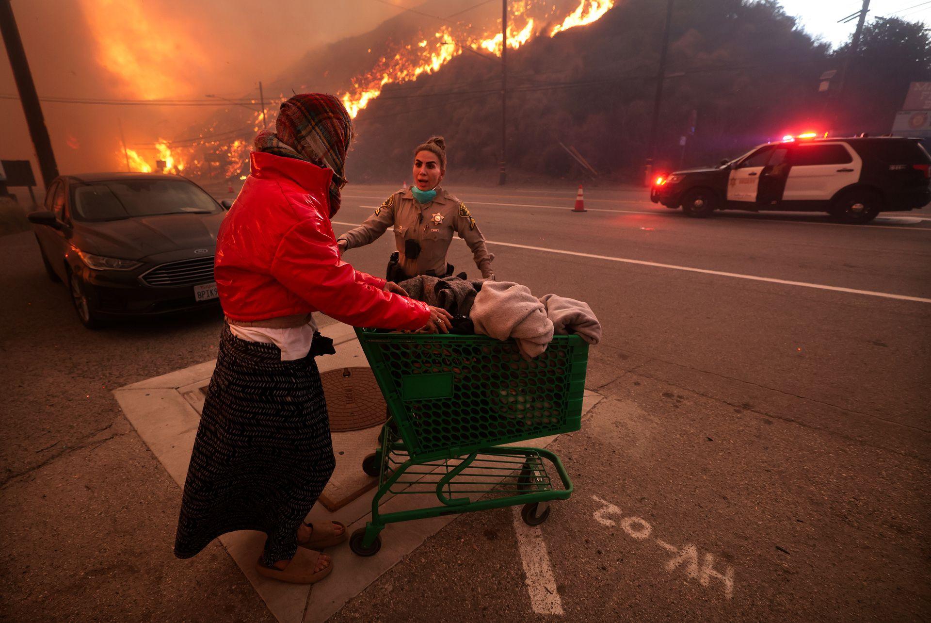 A police officer escorts a homeless woman pushing her belongings on a trolley, on a street with a police car and burning mountain in the background, in Topanga Canyon Blvd.