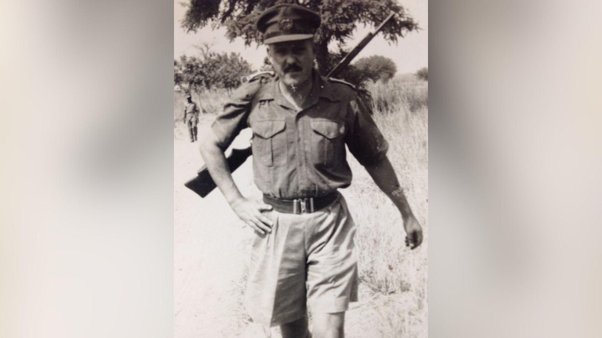 Jim Almonds, pictured in military issue shorts and short sleeves, is walking through grasslands. He has a rifle slung over his back.