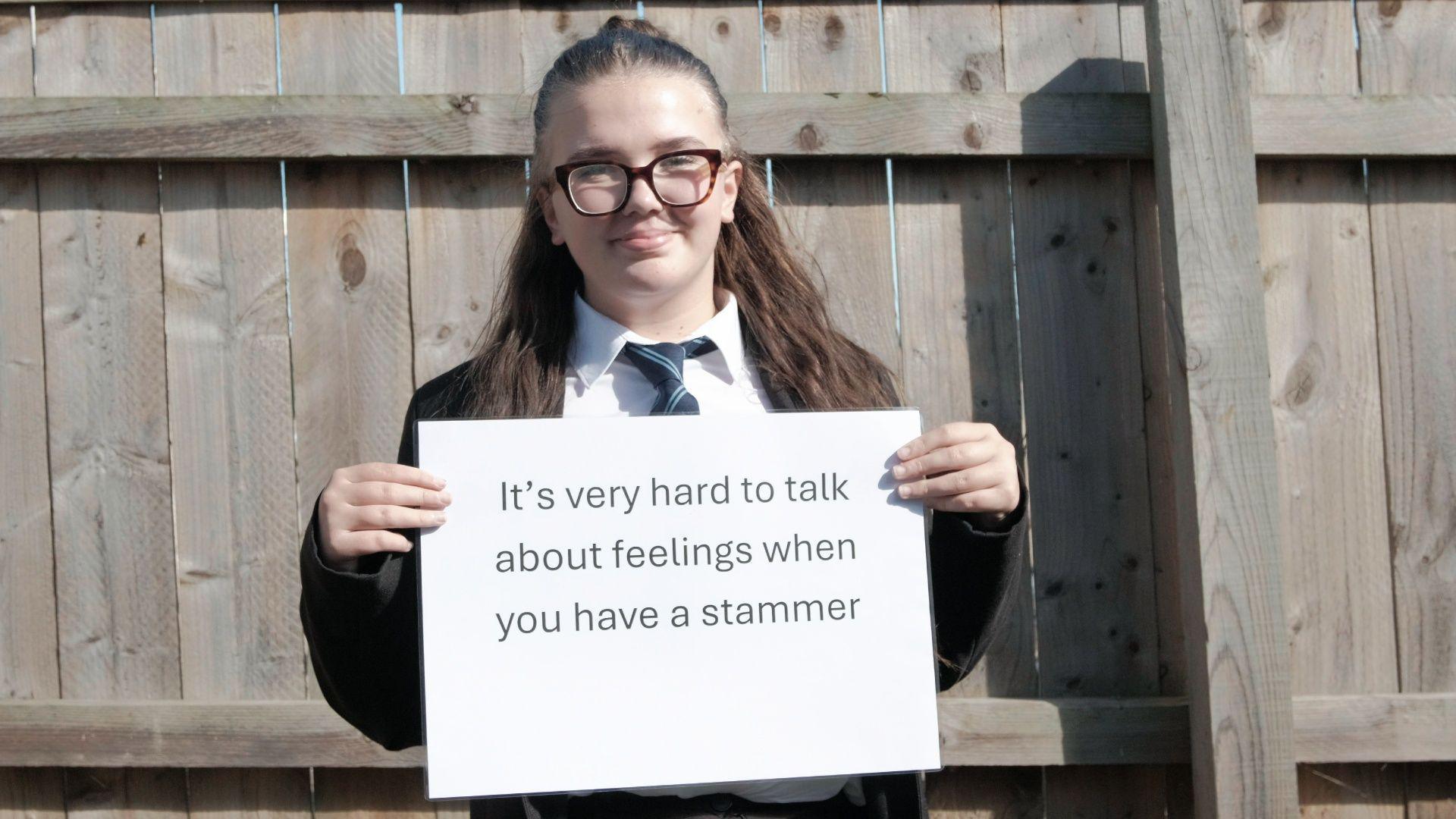 A teenage girl with tortoiseshell large glasses has brown hair half up half down and is smiling whilst holding a sign. The sign reads 'it's very hard to talk about feelings when you have a stammer'. She is standing in front of a wooden fence and is wearing a school uniform including and black and blue stripy tie.