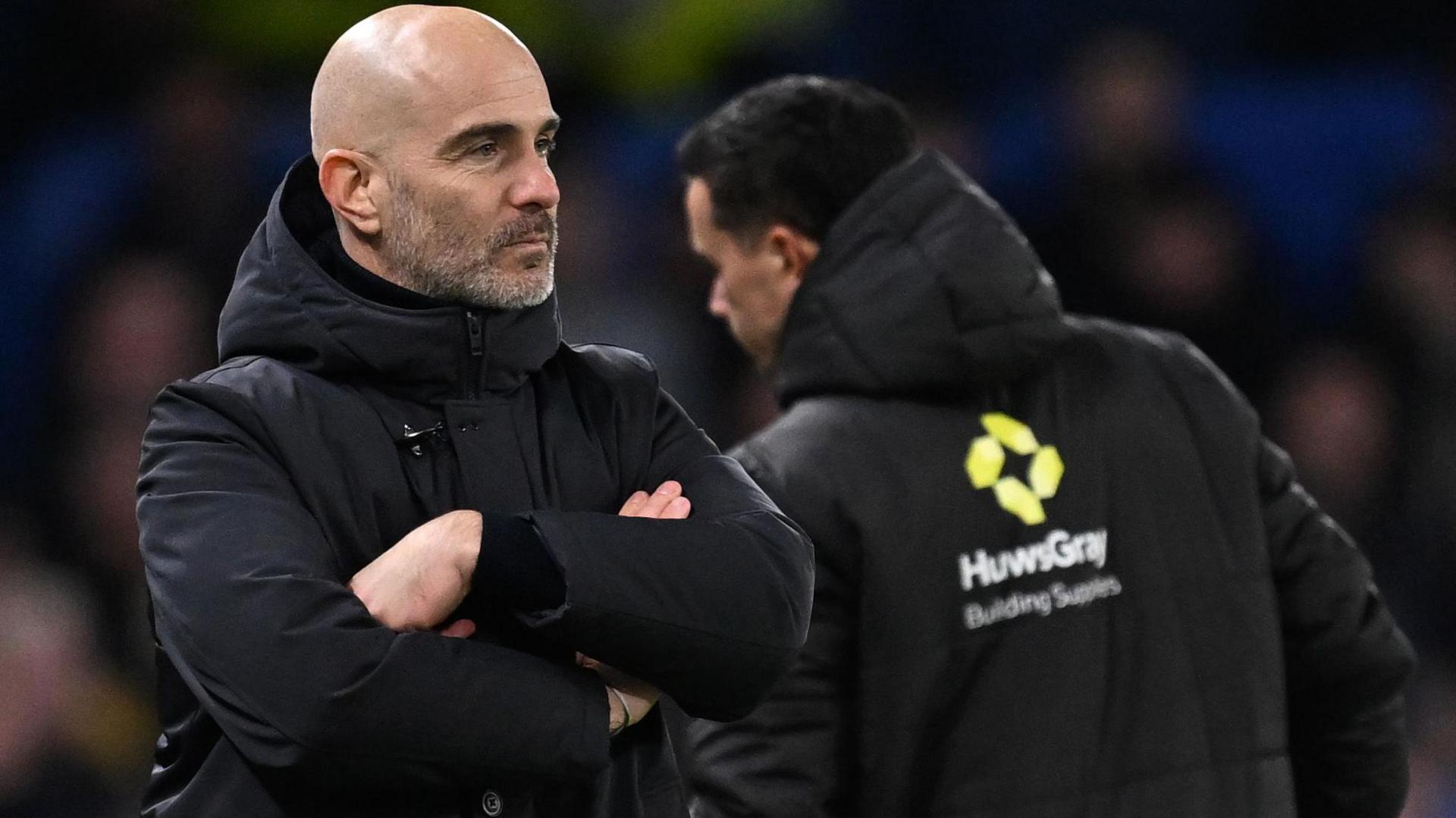 Enzo Maresca, wearing a black hooded coat and looking serious, stands with his arms folded on the touchline