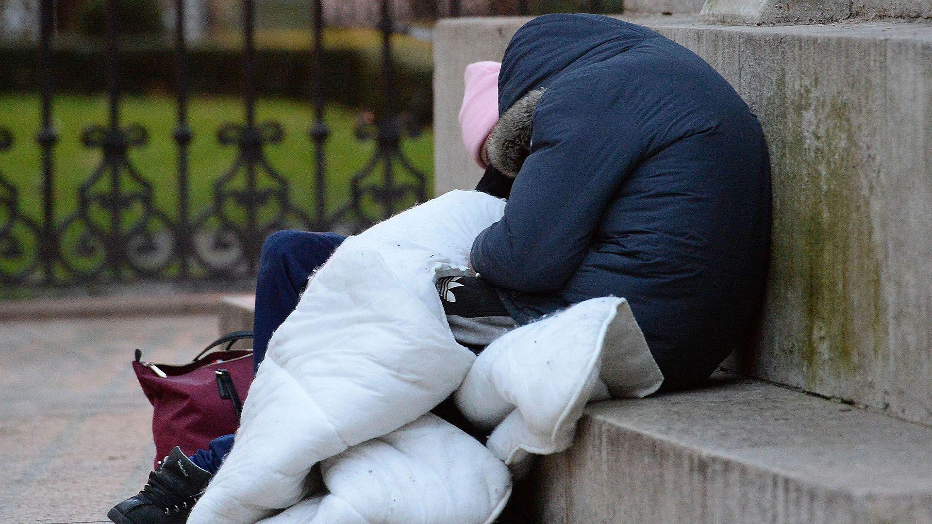 Two rough sleepers wearing coats and a blanket huddle together on a concrete step.