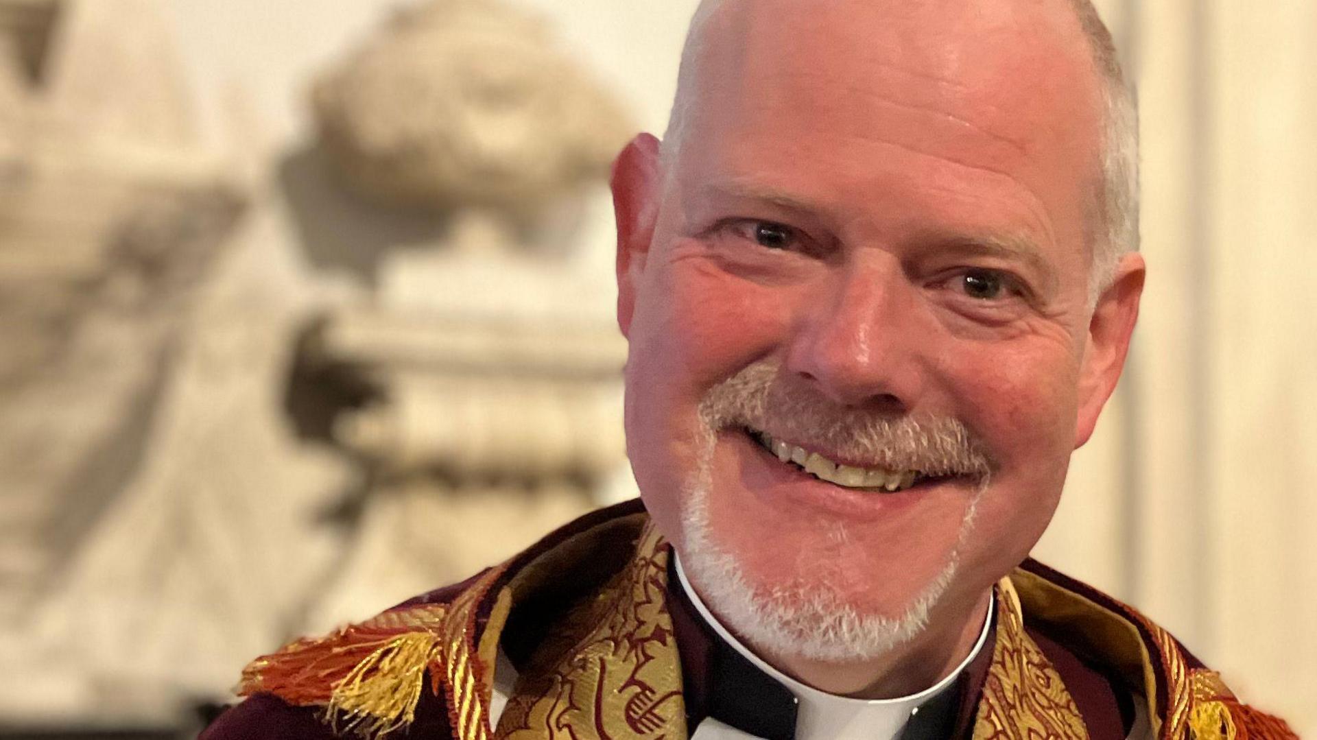 A man with grey hair and beard smile at the camera. He is wearing the religious attire of a Dean, including a dog collar, a red and gold stole and a dark coloured cape 