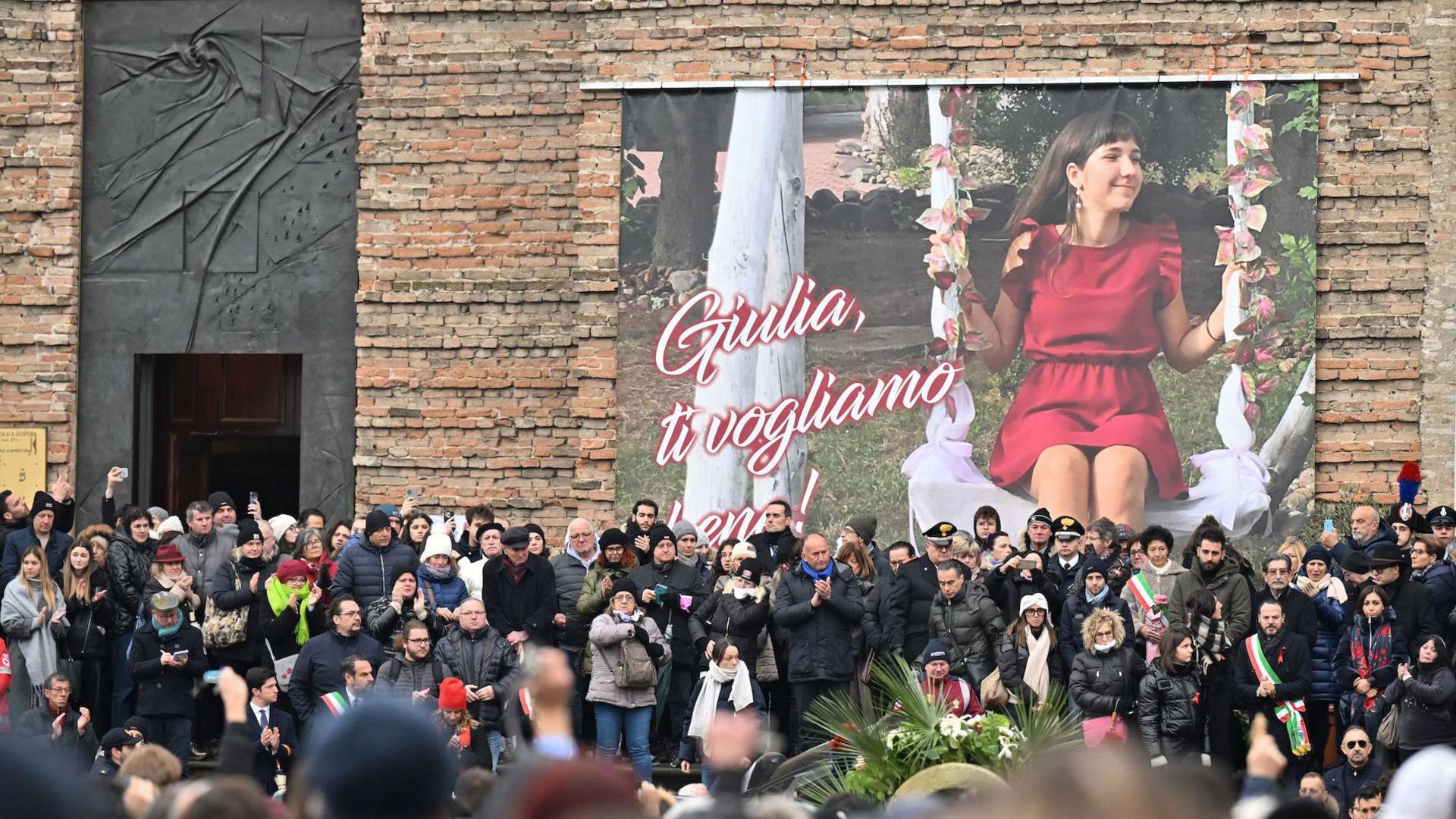 A large crowd of people outside the Basilica of Santa Giustina. On the wall of the basilica is a large poster several metres high showing Giulia Cecchettin in a red dress sitting on a swing.
