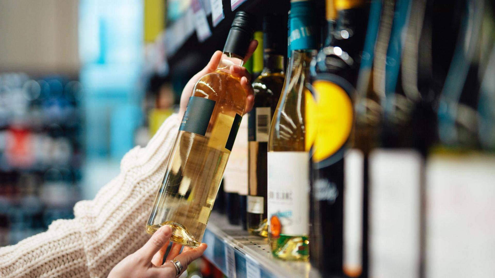 Stock picture of a woman's hand picking up a bottle of white wine from a supermarket shelf.