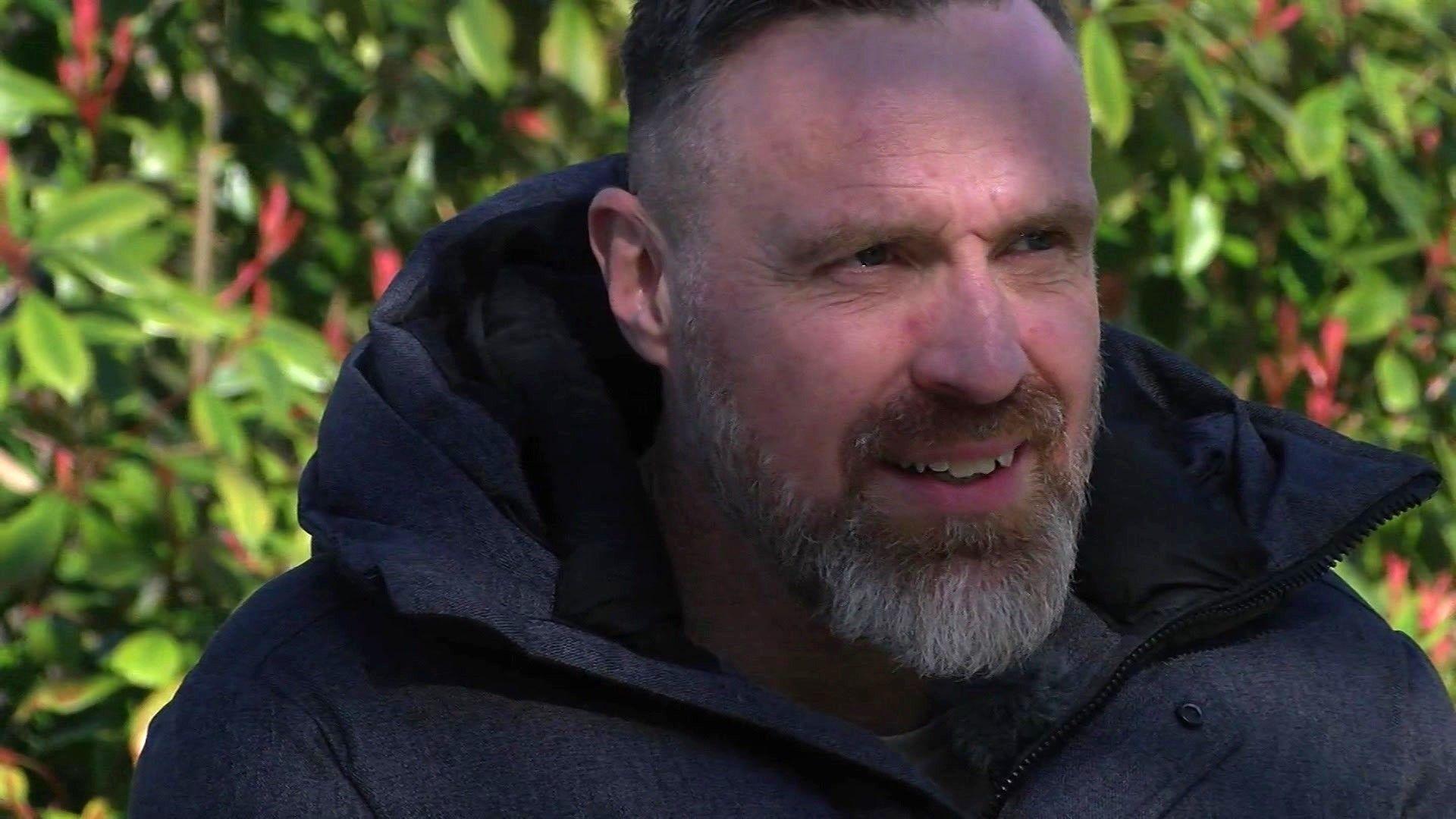 Allan, a man with short dark grey hair with a light grey beard, wearing a navy blue coat. Behind him is a green, leafy bush.