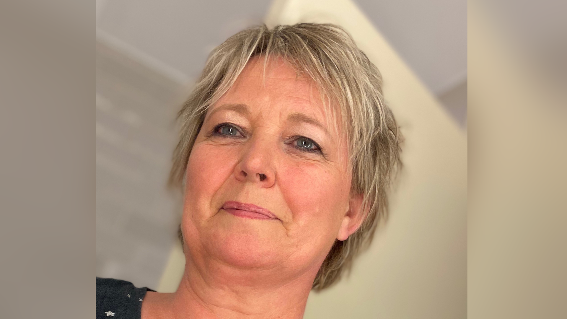 Alison has short blonde hair and blue eyes. She is looking to the left of the camera and smiling. She is stood against a plain cream coloured background.