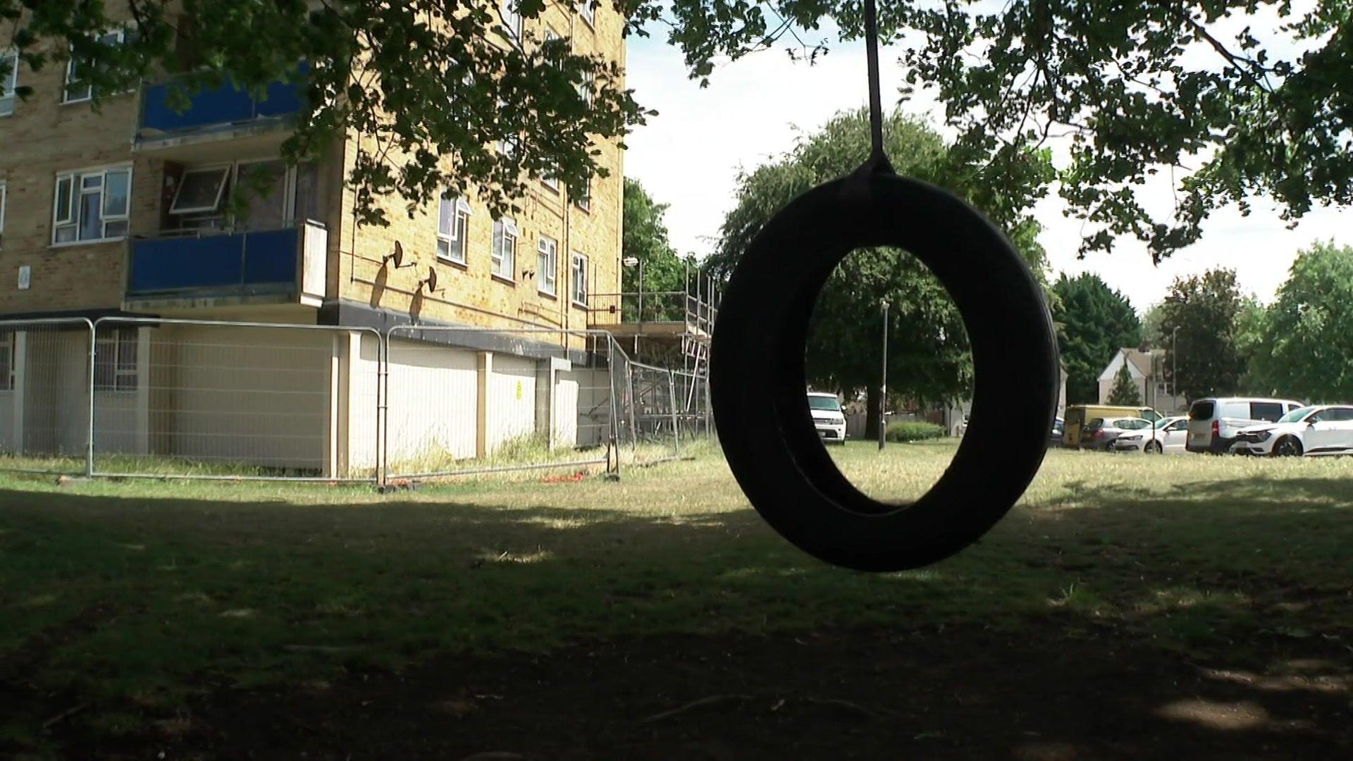 Close-up of a large tyre swing, tree branches hanging down, and behind the bottom of a tower block being repaired, with large metal fencing and scaffolding.