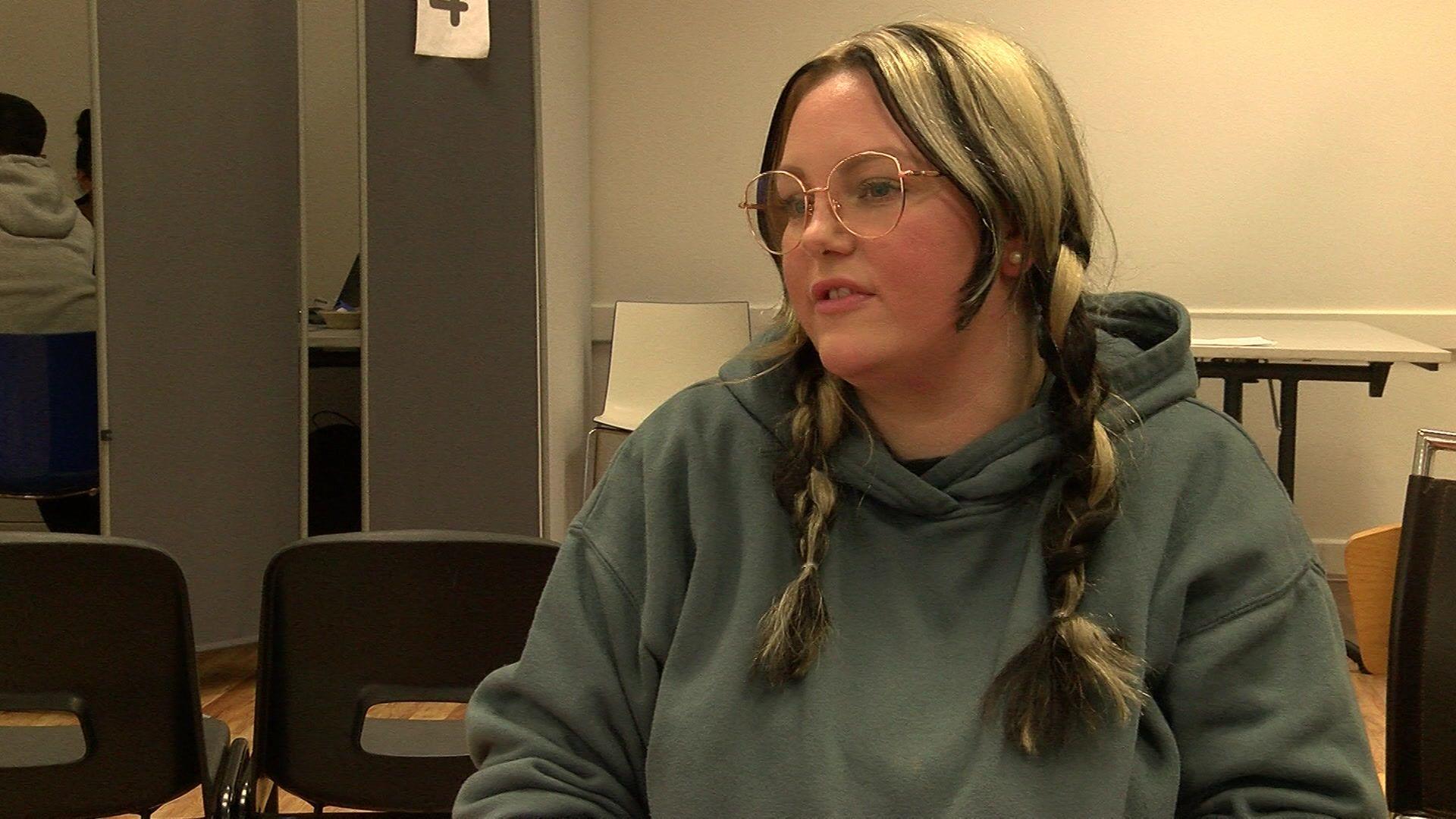 A young woman with dark blonde hair, worn in pigtails. She wears glasses and a green hooded top.