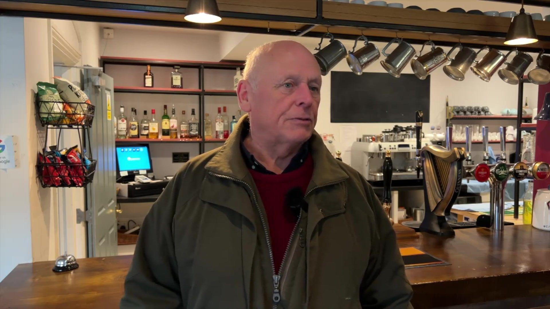 A bald man in a burgundy jumper and a forest green coat. He is standing in front of a pub bar.