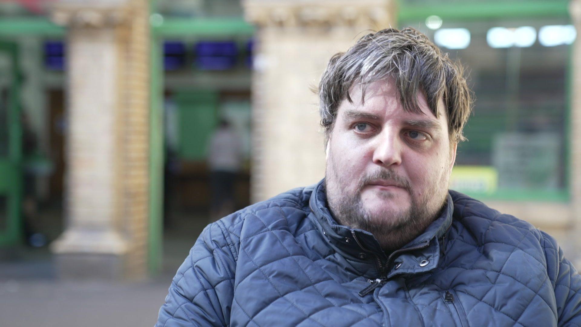 Nathan Rollinson is seen with short brown hair and a beard wearing a navy quilted jacket, pictured in front of a brick wall and green doorways.
