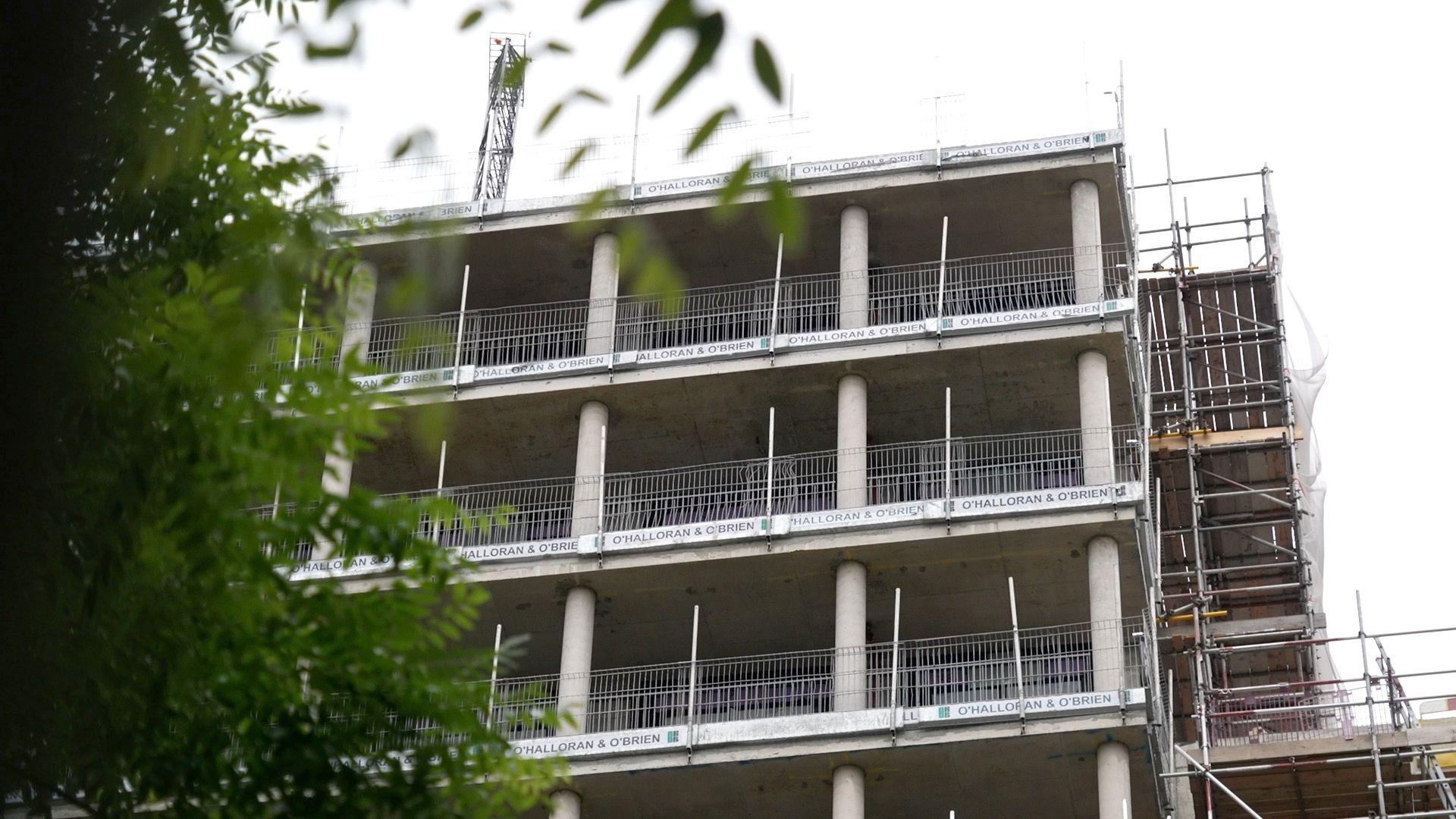 A housing development under construction. Concrete storeys are installed with wire safety netting at the side. Scaffolding goes up the right side of the building.