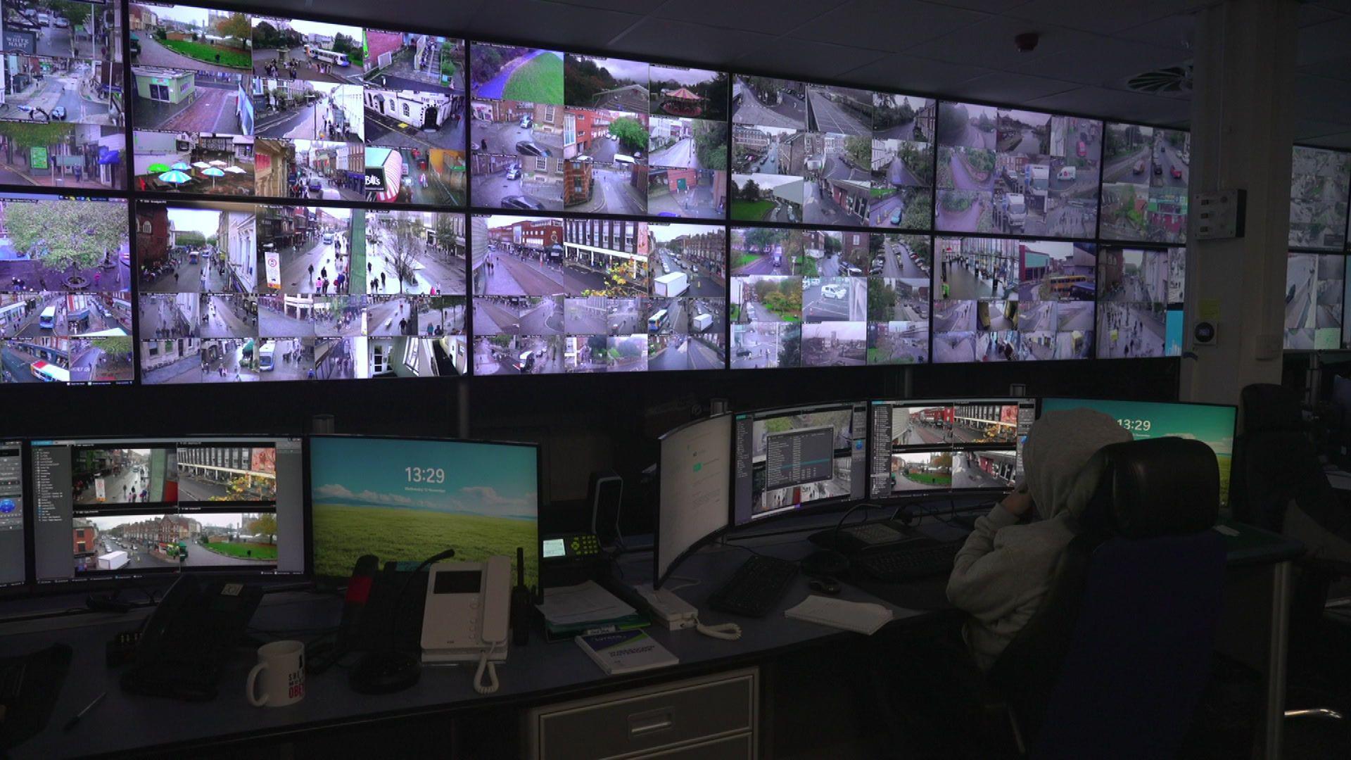 A wall filled with monitors which show CCTV footage from around Exeter. There are a few computers in the foreground.