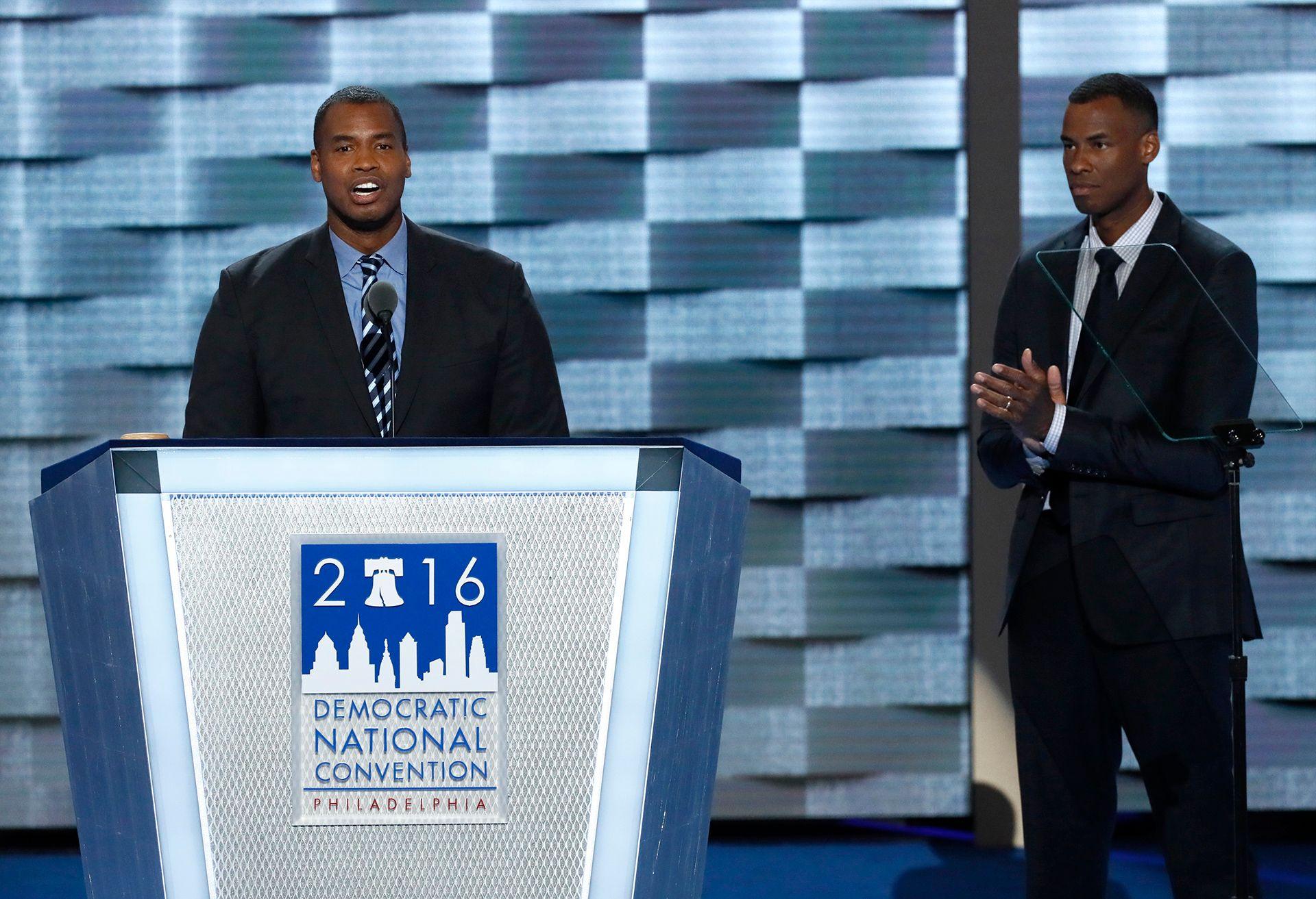 Collins speaks into a microphone while standing at a lecturn with a sign reading '2016 Democratic National Convention Philadelphia', while his brother Jarron applauds while standing onstage next to him.