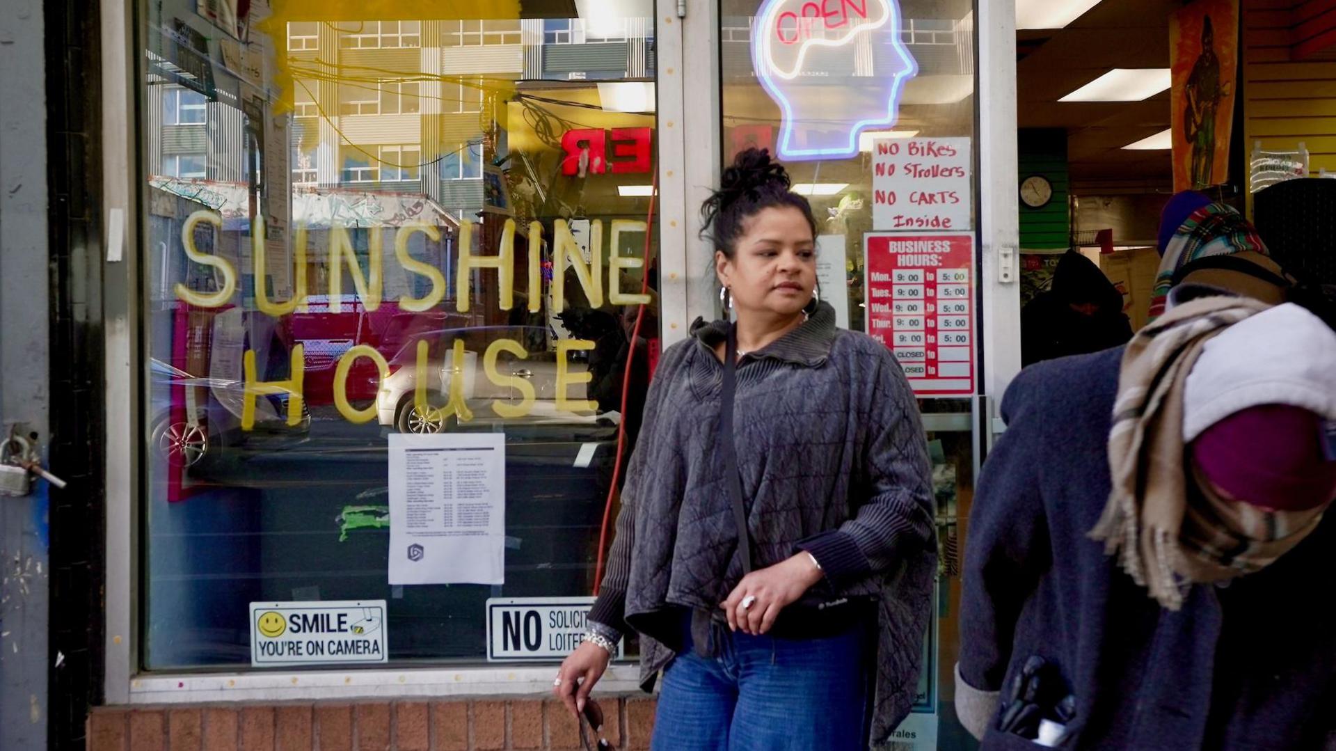 Rosalind Pichardo, a woman with her black her up in a bun, wearing a grey top and blue jeans, standing outside her drop-in centre which has "Sunshine House" written on the window. She looks towards the doorway as a stream of people enter the centre.