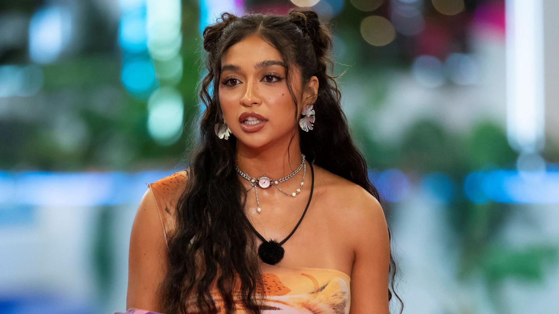 Cierra Ortega on Love Island USA. Cierra has long, wavy dark hair and brown eyes. She wears a patterned one-shouldered orange dress and a microphone around her neck.