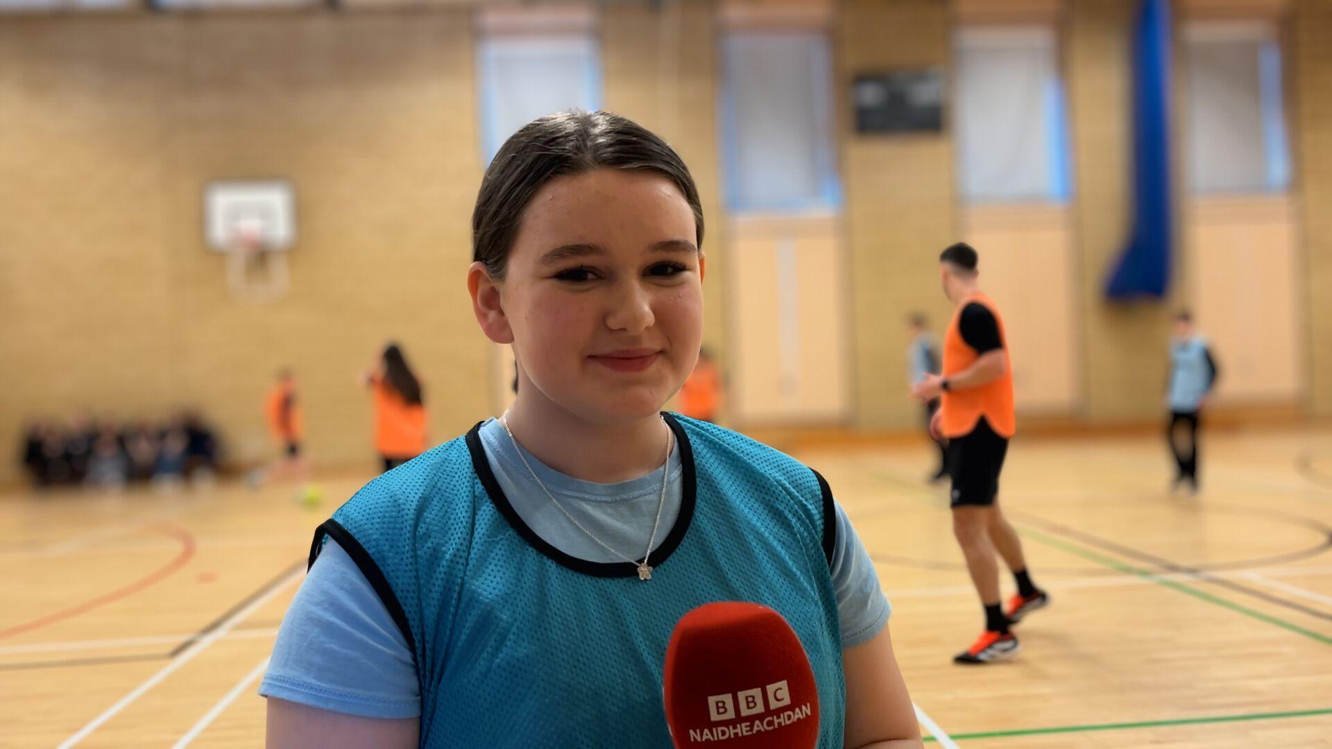 Kerry, sgoilear aig Sgoil Sir E Scott na seasamh san talla spòrs agus cuid de sgoilearan is cuid de phoilis a' cluiche ball-coise air a cùlaibh.