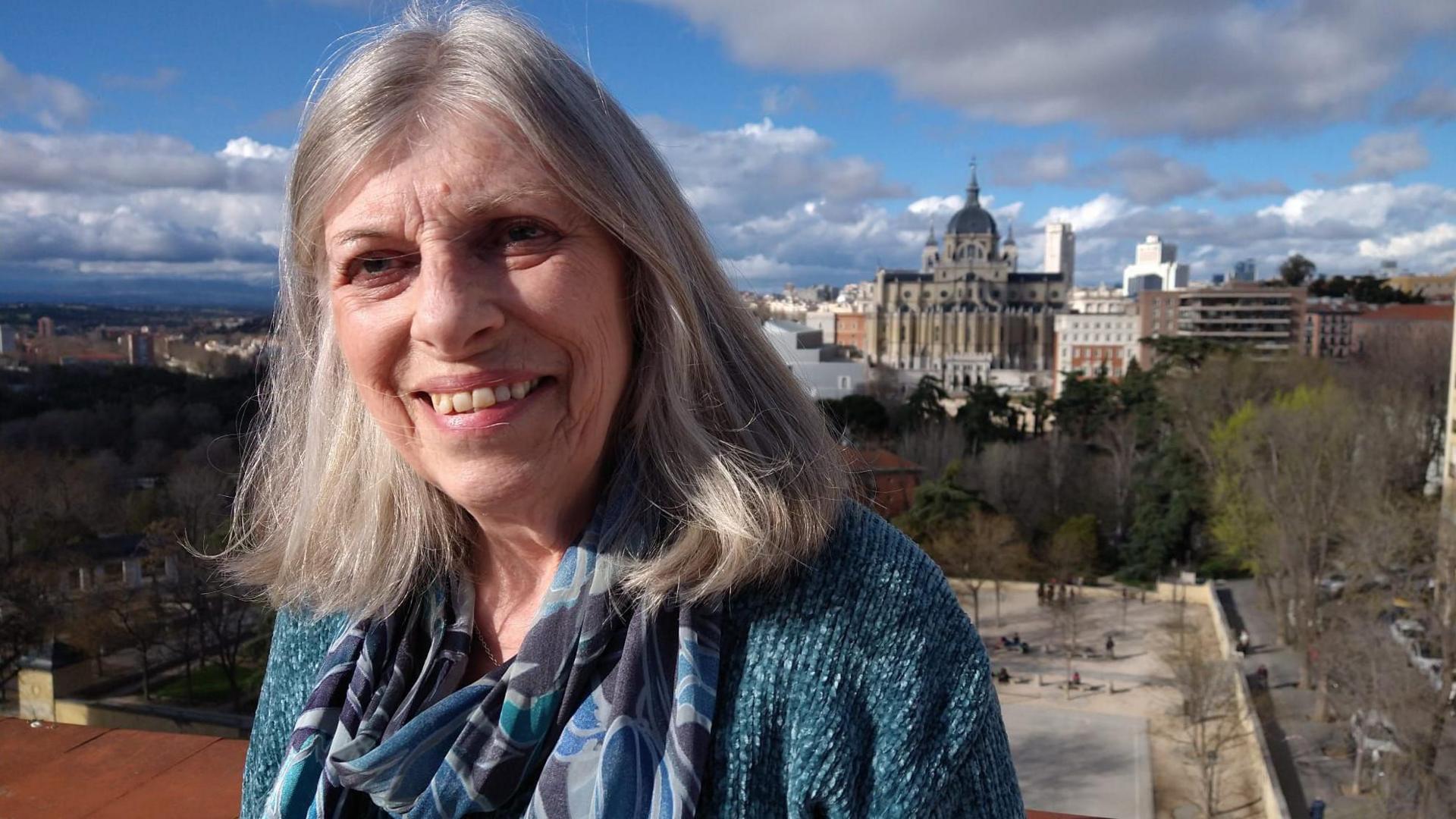 76-year-old Valerie Stacey smiles at the camera, her long, white hair is down and she's wearing a blue scarf. She is standing in a rooftop terrace overseeing the cathedral in Madrid.