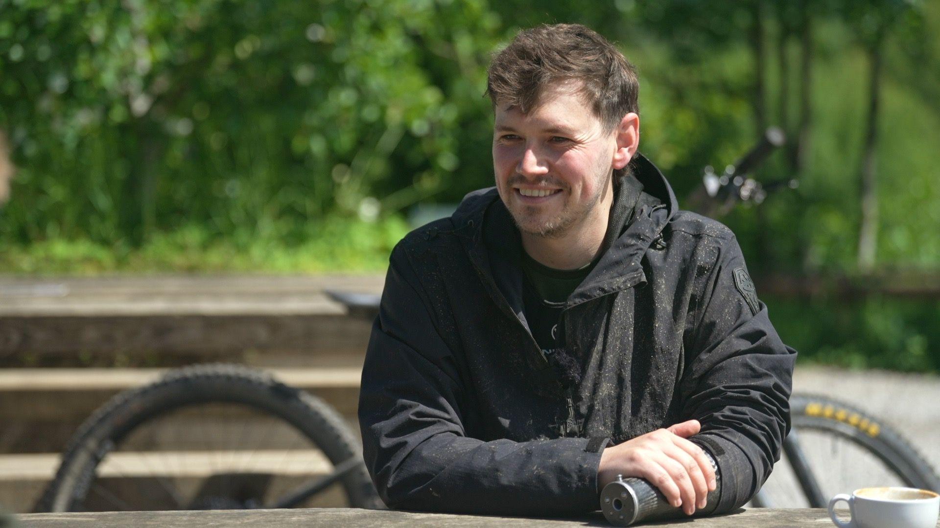 Morgan Jones wearing a black jacket and with a prosthetic left arm smiles at the camera, with a mountain bike in the background.