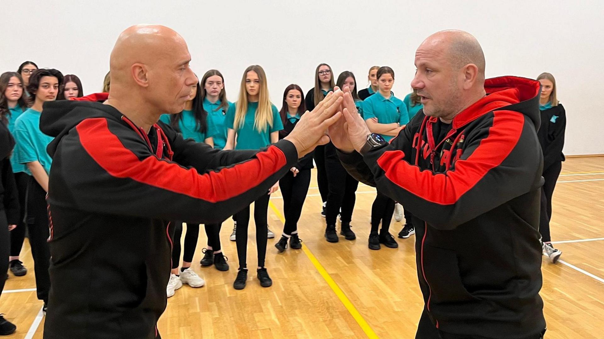 Two Krav Maga tutors face one another with arms outstretched and hands touching as they demonstrate a self-defence technique. Both men have shaven heads and wear black and red tracksuits. They are watched by about 15 teenage girls wearing aquamarine tops and black leggings. 