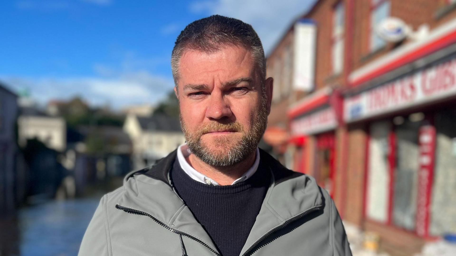 A grey haired man wears a grey waterproof jacket, black jumper and white shirt. He has a short brown beard. Over his right shoulder is an out-of-focus street with grey buildings. Over his left is a row of bricked properties, the one closest to him has a red shop front and glass windows. 