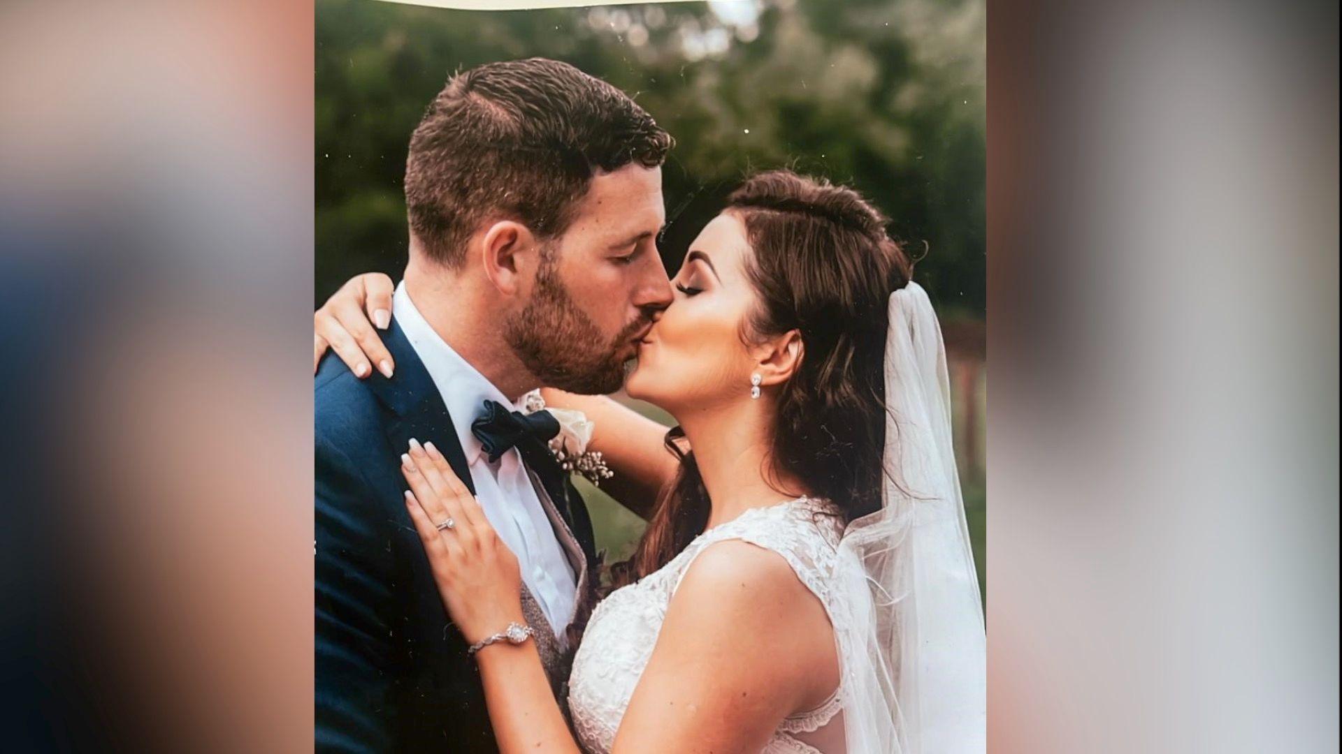 A couple kisses. He has brown short hair and she has long brown hair and wears a white sleeveless dress, silver watch and veil at the back of her head. 