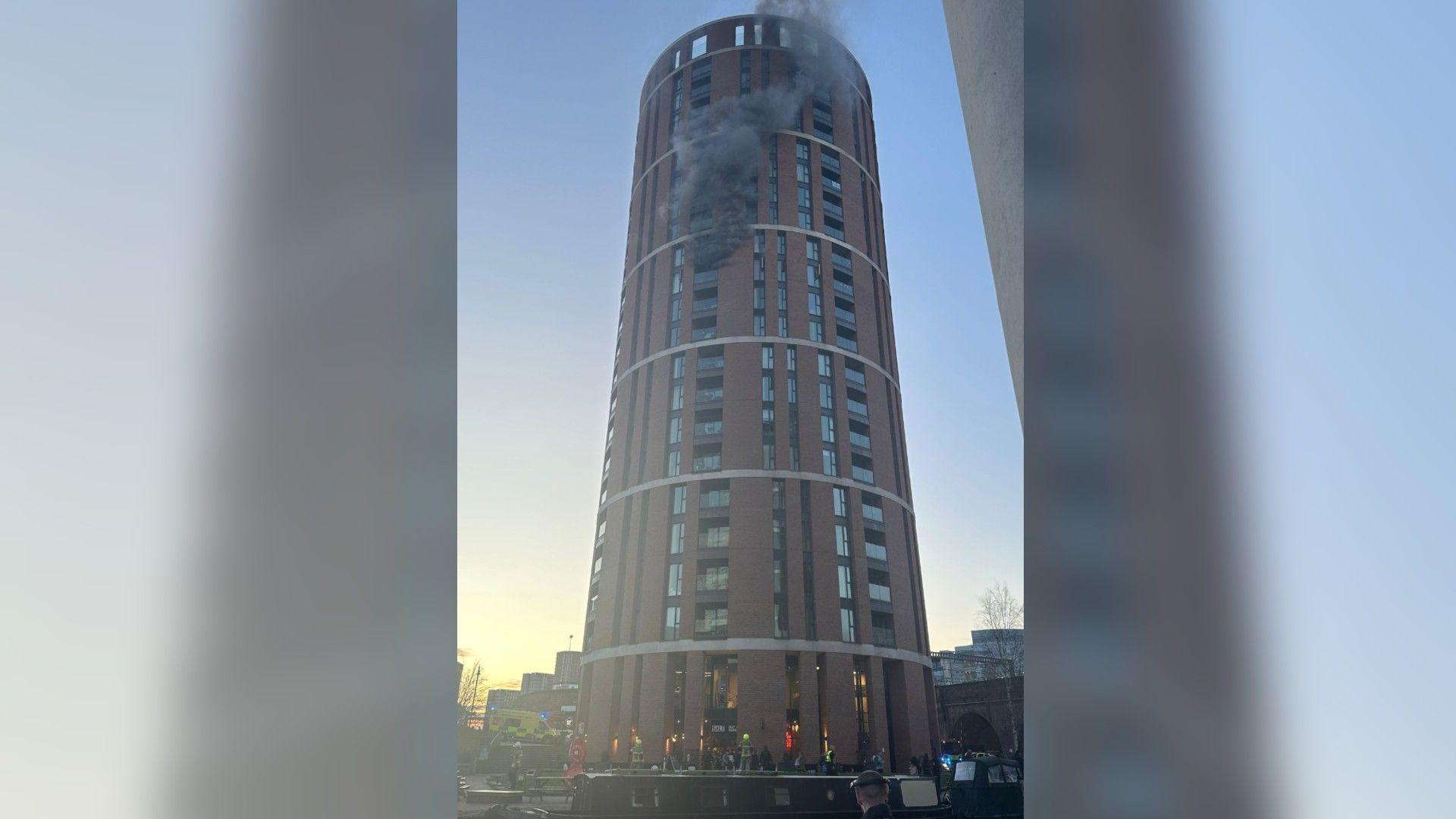 A high-rise, circular building with smoke billowing out of one of the upper floors. 