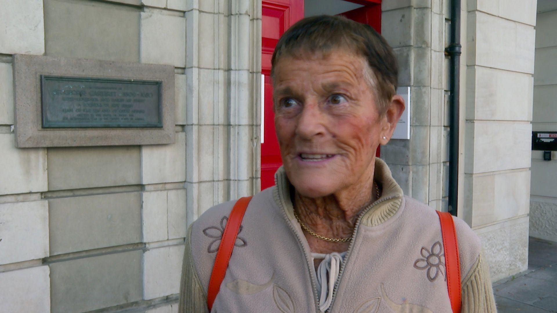 Judith Bennet looking to the left and wearing a light pink jumper with a flower print. She is wearing an orange rucksack and a gold necklace. She is stood outside of a white building which has red double doors and a grey/green plaque on the left.