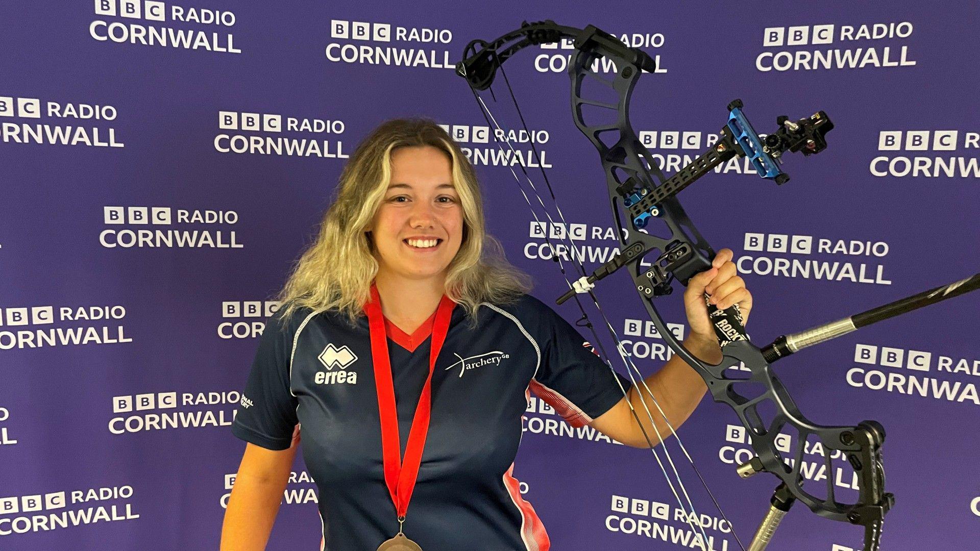 Chloe is smiling at the camera holding her compound bow up high. She is wearing her GB archery uniform and a bronze medal with a red ribbon. She has shoulder-length blonde hair. The purple backdrop is covered with repeated white BBC Radio Cornwall logos.