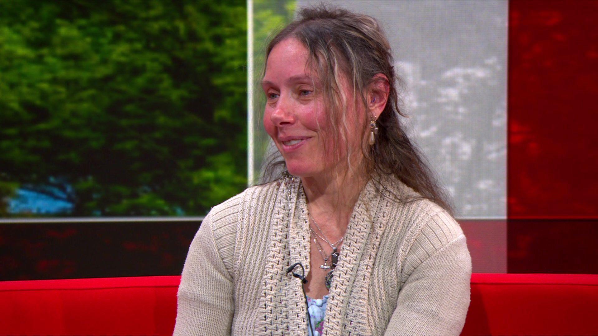 Ms Tindal in the BBC Spotlight studio wearing a beige cardigan. She has dark blonde hair and is sat on a red sofa with a tree background.