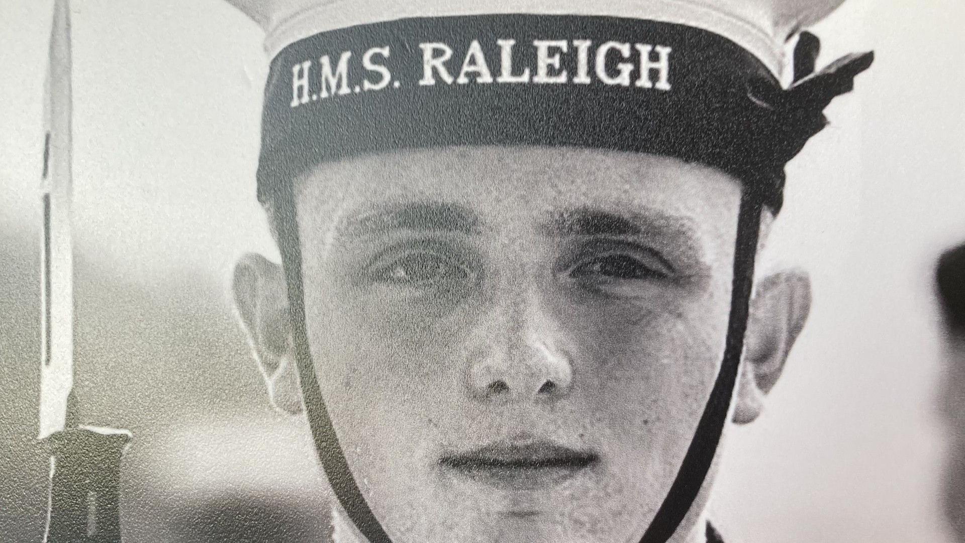 A black and white image of a young man in a navy cap with the word HMS RALEIGH on the cap. The top of a bayonet can be seen to the left of the photo.