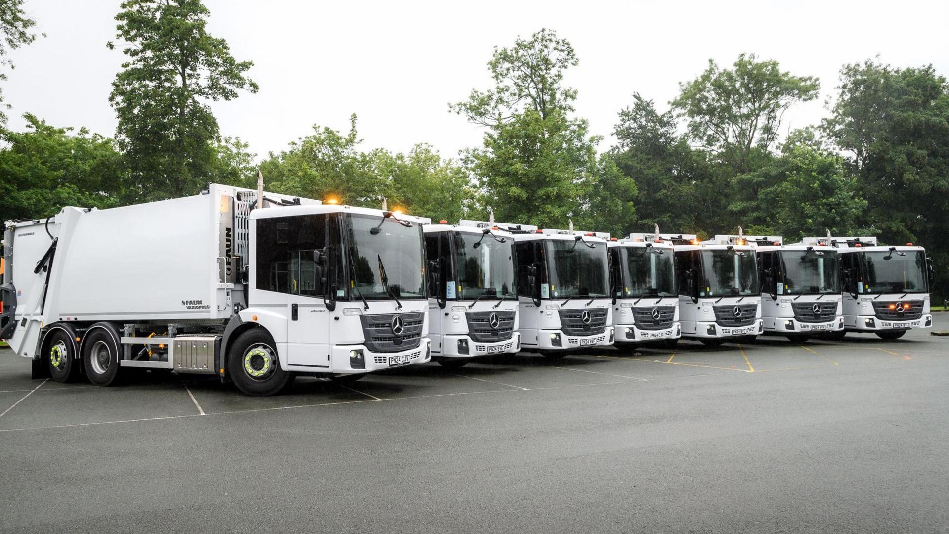 New fleet of bin lorries lined up in a row in a car park with trees behind. They are white with windows at the front. 