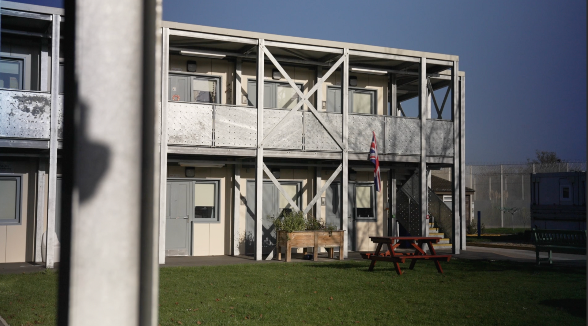 Part of HMP Send, a building is in the background behind grass and a picnic bench.